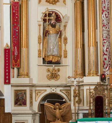 Zdjęcie nr 4: Całopostaciowa, drążona z tyłu figura św. Wojciecha stojącego w układzie frontalnym, w nieznacznym kontrapoście, na niewielkim, prostopadłościennym postumencie. Pociągłą twarz świętego charakteryzują sumarycznie opracowane rysy z szeroko rozstawionymi, lekko przymkniętymi oczami, zarumienione policzki i czubek nosa oraz długi, brązowy, skręcony zarost. Święty ubrany jest w strój pontyfikalny składający się ze złoconej alby, srebrzonej komży, złoconego mucetu oraz złoconej kapy zdobionej motywami roślinnymi z obszyciem z kaboszonami. Na głowie ma złocono-srebrzoną infułę z dekoracją z kaboszonów i opadającymi na ramiona srebrzonymi taśmami. Dłonie biskupa osłaniają czerwone rękawiczki, a stopy czerwone buty. W prawej ręce trzyma krzyż pastoralny, a lewej wiosło.