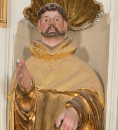 Zdjęcie nr 1: Całopostaciowa, drążona z tyłu figura św. Dominika stojącego w układzie frontalnym, w kontrapoście, na niewielkim, prostopadłościennym postumencie. Kwadratową w obrysie twarz cechują sumarycznie opracowane rysy z wydatnym nosem, niewielkimi, szeroko rozstawionymi oczami oraz podkreślonymi rumieńcem policzkami i czubkiem nosa oraz krótkim zarostem. Krótko przystrzyżone, skręcone włosy z wygoloną tonsurą odsłaniają duże, przylegające do głowy uszy. Święty ubrany jest w złocony habit dominikański ze szkaplerzem i kukullą, której połę podtrzymuje lewą ręką, prawą unosi w geście błogosławieństwa. Dłonie świętego są duże i szerokie o sumarycznym modelunku placów. U stóp świętego, po lewej stronie znajduje się srebrzona sylwetka leżącego psa.     