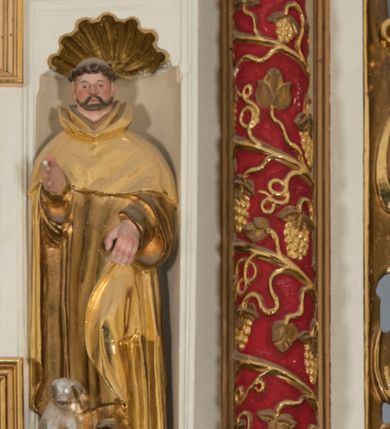 Zdjęcie nr 2: Całopostaciowa, drążona z tyłu figura św. Dominika stojącego w układzie frontalnym, w kontrapoście, na niewielkim, prostopadłościennym postumencie. Kwadratową w obrysie twarz cechują sumarycznie opracowane rysy z wydatnym nosem, niewielkimi, szeroko rozstawionymi oczami oraz podkreślonymi rumieńcem policzkami i czubkiem nosa oraz krótkim zarostem. Krótko przystrzyżone, skręcone włosy z wygoloną tonsurą odsłaniają duże, przylegające do głowy uszy. Święty ubrany jest w złocony habit dominikański ze szkaplerzem i kukullą, której połę podtrzymuje lewą ręką, prawą unosi w geście błogosławieństwa. Dłonie świętego są duże i szerokie o sumarycznym modelunku placów. U stóp świętego, po lewej stronie znajduje się srebrzona sylwetka leżącego psa.     