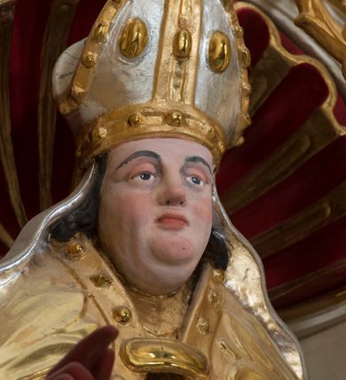 Zdjęcie nr 4: Całopostaciowa, drążona z tyłu figura św. Stanisława biskupa stojącego w układzie frontalnym, w nieznacznym kontrapoście, na niewielkim postumencie. Owalną, pociągłą twarz charakteryzują wyraziste, sumarycznie opracowane rysy z krótkim, lekko zadartym nosem, dużymi, szeroko rozstawionymi oczami o delikatnie przymkniętych powiekach, wyrazistymi łukami brwi oraz zarumienionymi policzkami, czubkiem nosa i wydatnym podbródkiem. Ciemnobrązowe, falowane włosy wspierają się na barkach. Św. Stanisław ubrany jest w strój pontyfikalny składający się ze złoconej alby, srebrzonej komży, złoconego mucetu i spiętej pod szyją, złoconej kapy zdobionej motywami floralnymi oraz obszyciem z kaboszonami. Dłonie biskupa osłaniają czerwone rękawiczki, stopy czerwone buty. Głowę wieńczy srebrzona i złocona infuła zdobiona aplikacjami w formie kaboszonów, której srebrzone taśmy opadają na ramiona świętego. Święty w lewej ręce trzyma pastorał, prawą unosi w geście błogosławieństwa. U jego stóp, po lewej stronie widoczne jest półpostaciowe przedstawienie Piotrowina wydostającego się z prostokątnego, srebrzonego sarkofagu. Sylwetkę mężczyzny osłania złocony płaszcz narzucony także na podniesioną do góry głowę. Twarz charakteryzują sumarycznie opracowane rysy ze spiczasto zakończonym nosem i wydatnymi wąsami. Duże, szerokie dłonie złożone są w geście modlitewnym.  