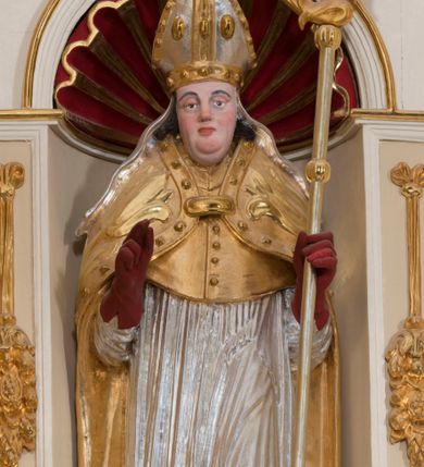 Zdjęcie nr 1: Całopostaciowa, drążona z tyłu figura św. Stanisława biskupa stojącego w układzie frontalnym, w nieznacznym kontrapoście, na niewielkim postumencie. Owalną, pociągłą twarz charakteryzują wyraziste, sumarycznie opracowane rysy z krótkim, lekko zadartym nosem, dużymi, szeroko rozstawionymi oczami o delikatnie przymkniętych powiekach, wyrazistymi łukami brwi oraz zarumienionymi policzkami, czubkiem nosa i wydatnym podbródkiem. Ciemnobrązowe, falowane włosy wspierają się na barkach. Św. Stanisław ubrany jest w strój pontyfikalny składający się ze złoconej alby, srebrzonej komży, złoconego mucetu i spiętej pod szyją, złoconej kapy zdobionej motywami floralnymi oraz obszyciem z kaboszonami. Dłonie biskupa osłaniają czerwone rękawiczki, stopy czerwone buty. Głowę wieńczy srebrzona i złocona infuła zdobiona aplikacjami w formie kaboszonów, której srebrzone taśmy opadają na ramiona świętego. Święty w lewej ręce trzyma pastorał, prawą unosi w geście błogosławieństwa. U jego stóp, po lewej stronie widoczne jest półpostaciowe przedstawienie Piotrowina wydostającego się z prostokątnego, srebrzonego sarkofagu. Sylwetkę mężczyzny osłania złocony płaszcz narzucony także na podniesioną do góry głowę. Twarz charakteryzują sumarycznie opracowane rysy ze spiczasto zakończonym nosem i wydatnymi wąsami. Duże, szerokie dłonie złożone są w geście modlitewnym.  
