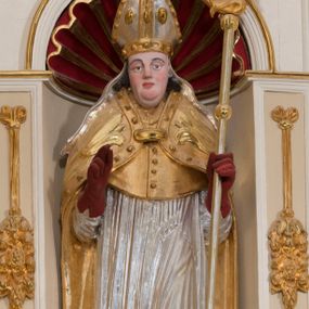 Zdjęcie nr 1: Całopostaciowa, drążona z tyłu figura św. Stanisława biskupa stojącego w układzie frontalnym, w nieznacznym kontrapoście, na niewielkim postumencie. Owalną, pociągłą twarz charakteryzują wyraziste, sumarycznie opracowane rysy z krótkim, lekko zadartym nosem, dużymi, szeroko rozstawionymi oczami o delikatnie przymkniętych powiekach, wyrazistymi łukami brwi oraz zarumienionymi policzkami, czubkiem nosa i wydatnym podbródkiem. Ciemnobrązowe, falowane włosy wspierają się na barkach. Św. Stanisław ubrany jest w strój pontyfikalny składający się ze złoconej alby, srebrzonej komży, złoconego mucetu i spiętej pod szyją, złoconej kapy zdobionej motywami floralnymi oraz obszyciem z kaboszonami. Dłonie biskupa osłaniają czerwone rękawiczki, stopy czerwone buty. Głowę wieńczy srebrzona i złocona infuła zdobiona aplikacjami w formie kaboszonów, której srebrzone taśmy opadają na ramiona świętego. Święty w lewej ręce trzyma pastorał, prawą unosi w geście błogosławieństwa. U jego stóp, po lewej stronie widoczne jest półpostaciowe przedstawienie Piotrowina wydostającego się z prostokątnego, srebrzonego sarkofagu. Sylwetkę mężczyzny osłania złocony płaszcz narzucony także na podniesioną do góry głowę. Twarz charakteryzują sumarycznie opracowane rysy ze spiczasto zakończonym nosem i wydatnymi wąsami. Duże, szerokie dłonie złożone są w geście modlitewnym.  