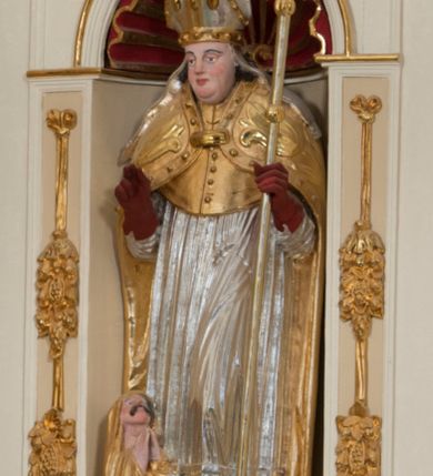 Zdjęcie nr 2: Całopostaciowa, drążona z tyłu figura św. Stanisława biskupa stojącego w układzie frontalnym, w nieznacznym kontrapoście, na niewielkim postumencie. Owalną, pociągłą twarz charakteryzują wyraziste, sumarycznie opracowane rysy z krótkim, lekko zadartym nosem, dużymi, szeroko rozstawionymi oczami o delikatnie przymkniętych powiekach, wyrazistymi łukami brwi oraz zarumienionymi policzkami, czubkiem nosa i wydatnym podbródkiem. Ciemnobrązowe, falowane włosy wspierają się na barkach. Św. Stanisław ubrany jest w strój pontyfikalny składający się ze złoconej alby, srebrzonej komży, złoconego mucetu i spiętej pod szyją, złoconej kapy zdobionej motywami floralnymi oraz obszyciem z kaboszonami. Dłonie biskupa osłaniają czerwone rękawiczki, stopy czerwone buty. Głowę wieńczy srebrzona i złocona infuła zdobiona aplikacjami w formie kaboszonów, której srebrzone taśmy opadają na ramiona świętego. Święty w lewej ręce trzyma pastorał, prawą unosi w geście błogosławieństwa. U jego stóp, po lewej stronie widoczne jest półpostaciowe przedstawienie Piotrowina wydostającego się z prostokątnego, srebrzonego sarkofagu. Sylwetkę mężczyzny osłania złocony płaszcz narzucony także na podniesioną do góry głowę. Twarz charakteryzują sumarycznie opracowane rysy ze spiczasto zakończonym nosem i wydatnymi wąsami. Duże, szerokie dłonie złożone są w geście modlitewnym.  