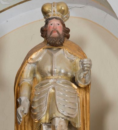 Zdjęcie nr 1: Całopostaciowa, drążona z tyłu rzeźba przedstawia św. Wacława stojącego w ujęciu frontalnym, w lekkim kontrapoście, na niewielkim, prostopadłościennym postumencie. Owalną w obrysie twarz mężczyzny charakteryzują sumarycznie opracowane rysy z prostym, spiczasto zakończonym nosem, wydatnymi kośćmi policzkowymi podkreślonymi rumieńcem, szeroko otwartymi oczami o szeroko rozstawionych, opadających łukach brwi oraz krótki, brązowy zarost. Po bokach twarz ujmują wydatne pukle półdługich, brązowych włosów. Święty ubrany jest w srebrzoną i złoconą rycerską zbroję płytową, plecy i ramiona osłania spięty pod szyją, długi, złocony płaszcz. Na głowie ma złoconą i srebrzoną mitrę książęcą. Prawa ręka opuszczona jest wzdłuż ciała, w lewej, zgiętej w łokciu pierwotnie znajdował się niezachowany atrybut świętego.  