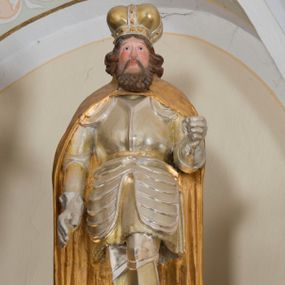 Zdjęcie nr 1: Całopostaciowa, drążona z tyłu rzeźba przedstawia św. Wacława stojącego w ujęciu frontalnym, w lekkim kontrapoście, na niewielkim, prostopadłościennym postumencie. Owalną w obrysie twarz mężczyzny charakteryzują sumarycznie opracowane rysy z prostym, spiczasto zakończonym nosem, wydatnymi kośćmi policzkowymi podkreślonymi rumieńcem, szeroko otwartymi oczami o szeroko rozstawionych, opadających łukach brwi oraz krótki, brązowy zarost. Po bokach twarz ujmują wydatne pukle półdługich, brązowych włosów. Święty ubrany jest w srebrzoną i złoconą rycerską zbroję płytową, plecy i ramiona osłania spięty pod szyją, długi, złocony płaszcz. Na głowie ma złoconą i srebrzoną mitrę książęcą. Prawa ręka opuszczona jest wzdłuż ciała, w lewej, zgiętej w łokciu pierwotnie znajdował się niezachowany atrybut świętego.  