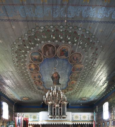 Zdjęcie nr 2: Ściany kościoła pokrywa dekoracja dywanowa, której w partiach stropu towarzyszą przedstawienia figuralne. W prezbiterium wykonano wyraźnie zaakcentowany fryz podstropowy, który od fasety oddziela profilowany i ornamentalnie malowany gzyms. Fryz na baldoniebieskim tle pokryto naprzemiennie rozmieszczonymi uskrzydlonymi główkami aniołów z niskimi wazonami z rozłożonymi trzema gałązkami kwitnących lilii. Poniżej barwnej bordiury oddzielającej fryz od pozostałych partii ścian, wykonano zgeometryzowaną dekorację utworzoną z powtarzających się wielobocznych pól, w których znalazł się stylizowany motyw tego samego kwiatu. Fasetę w prezbiterium pokryto naprzemiennie, rytmicznie rozmieszczonymi stylizowanymi kwiatami na łodydze i bez łodygi, które obwiedziono migdałowymi otokami tworzą ornamentalny pas zamknięty u góry falującą, wąską bordiurą. Dekorację bladoniebieskiego stropu prezbiterium tworzy poprowadzona przy krawędziach srebrno-niebieska arkatura. W centrum na niebieskim tle usianym złotymi gwiazdkami wyobrażono zasiadającą na obłokach Trójcę Świętą obramienioną pofalowaną, wielobarwną bordiurą otoczoną stylizowaną, srebrną wicią akantową. Dekoracyjną wić przedzielono parą uskrzydlonych główek anielskich oraz czterema stylizowanymi pąkami fantazyjnych kwiatów. Frontalnie ujęty Bóg Ojciec został wyobrażony jako siwobrody starzec o bujnych siwych włosach. W prawej ręce trzyma złote berło, lewą zaś wyciąga przed siebie. Bóg Ojciec prawą nogę opiera o niebieski glob, natomiast lewą, wyprostowaną wspiera się o obłoki. Jego twarz charakteryzuje jasna karnacja, szeroko otwarte oczy, wydatny nos i usta. Wokół głowy widać trójkąt. Obok Boga Ojca z lewej strony siedzi Chrystus, który obie nogi wspiera na globie. Zbawiciel trzymający oburącz na wysokości ramienia drewniany krzyż został przedstawiony w trzech czwartych w prawo. Jest to dojrzały mężczyzna o pociągłej twarzy okolonej brązowymi, długimi włosami spływającymi na ramiona oraz brodą. Ma jasną karnację, prosty nos i wydatne usta. Jego melancholijne spojrzenie jest skierowane w bok. Nagi tors okrywa bladoszkarłatny płaszcz odsłaniający prawą nogę. Na dłoniach i stopach widoczne są rany po gwoździach. Głowę Jezusa okala świetlisty nimb. Powyżej obu postaci unosi się gołębica Ducha Świętego w promienistym otoku. Niebieski glob na tle obłoków unoszą dwa aniołki w przepaskach. W nawie kościoła dekorację tworzą trzy różnej szerokości poziome pasy ornamentalne ułożone ponad jednolitą, szarą lamperią. Najszerszy pas pokryto na jasnym tle zgeometryzowanym ornamentem utworzonym z ciemnocynobrowych, migdałowo kształtowanych pól naśladujących wić roślinną. Powyżej rozmieszczono rytmicznie ułożone stylizowane krzyże równoramienne w fantazyjnych, wielobarwnych obramieniach. Najwyższy pas, odpowiadający fryzowi podstropowemu w prezbiterium pokryto wicią winnej latorośli z wplecionymi pękami kłosów. Tą część dekoracji zamyka profilowany gzyms ozdobiony wielobarwnym ornamentem zgeomertyzowanym. Rozpościerającą się ponad nim fasetę ozdobiono rytmicznie rozmieszczonym białym kwiatem na łodydze naśladujący w kształcie grzebień wodny. Dekorację bladoniebieskiego stropu artykułowano za pomocą srebrnej arkatury poprowadzonej przy jego krawędziach oraz ornamentalnych pasów utworzonych z motywów floralnych i geometrycznych, tworzących czworoboczną ramę dla centralnej kompozycji figuralnej. W narożach tego obramienia umieszczono medaliony ukazujące w popiersiach czterech ewangelistów malowanych na blasze miedzianej. Medaliony otaczają stylizowane gałązki dębowe z żołędziami. Każdego ewangelistę identyfikuje jego atrybut. W centrum stropu na bladozielonym tle usianym srebrnymi gwiazdkami namalowano wydłużone owalne obramienie utworzone z szerokiego wieńca różanego o bardzo drobnych kwiatach różanych i listkach. W jego centrum na tle bladoniebieskich obłoków ze złotymi gwiazdkami, bezpośrednio na deskach stropu, przedstawiono stojącą Madonnę z Dzieciątkiem, która depcze węża. Dodatkowo postać Marii zdaje się stać na rozpiętym różańcu. Maria ujęta frontalnie w kontrapoście obiema rękami podtrzymuje na prawym ramieniu Dzieciątko. Ma pociągłą twarz o jasnej karnacji, prostym nosie i wąskich ustach. Spojrzenie jej przymkniętych oczu jest skierowane w dół. Na głowie ma chustę szczelnie okrywającą jej włosy oraz złotą koronę o niskich sterczynach. Madonna ubrana jest w bladoczerwoną suknię odsłaniającą bosą stopę, którą depcze węża. Spod sukni na piersi widać białą koszulę. Na ramionach ma błękitny płaszcz podbity jasną ochrą, spięty na piersi podłużną, złotą klamrą. Trzymane przez nią Dzieciątko ma rozłożone ręce z uniesionymi dłońmi. Jego głowa ujęta jest w trzech czwartych w prawo. Otaczają ją jasno brązowe, krótkie włosy. Mały Jezus ubrany jest w jasną tunikę odsłaniającą jego bose stopy. Wokół Matki Boskiej rozmieszczono owalne medaliony z blachy miedzianej malowane w piętnaście tajemnic Różańca świętego. Ich narracja rozpoczyna się nad głową Madonny. Każda ze scen oparta jest o kilkuosobową kompozycję. Idąc w lewo namalowano: Zwiastowanie Marii, Nawiedzenie św. Elżbiety, Boże Narodzenie, Ofiarowanie w świątyni, Znalezienie w świątyni, Modlitwa w Ogrójcu, Biczowanie, Cierniem ukoronowanie, Niesienie krzyża, Ukrzyżowanie, Zmartwychwstanie, Wniebowstąpienie, Zesłanie Ducha Świętego, Wniebowzięcie Marii, Ukoronowanie Marii w niebie. Pomiędzy medalionami namalowano białą i czerwoną różę. Jasny parapet chóru muzycznego udekorowano naprzemiennie rozmieszczonymi rytmicznie stylizowanymi łodygami zakończonymi białymi pąkami i krzyżami równoramiennymi wpisanymi w koło. W aneksie południowym namalowano na ścianie kazanie św. Franciszka z Asyżu do ptaków. Kompozycja uzyskała kształt wydłużonego, stojącego prostokąta zamkniętego półkoliście i obwiedzionego falującą bordiurą z motywem łączących się krzyżyków. W jej centrum wyobrażono siedzącego na skalnym podłożu św. Franciszka ujętego w trzech czwartych w lewo. Święty prawą rękę unosi w geście mowy lewą zaś swobodnie trzyma na kolanach. Jego widoczną z profilu twarz o jasnej karnacji i prostym nosie okala siwiejąca broda i włosy z wyciętą tonsurą. Ubrany jest w brązowy habit przewiązany sznurem. Przed Franciszkiem przedstawiono stadko słuchających świetego ptaków. W prawym rogu kompozycji namalowano drewniane taczki. Z tyłu za świętym na tle skał rośnie drzewo. Inne drzewo wyobrażono z lewej strony, nieco z tyłu, na tle nieba pokrytego baldoniebieskimi obłokami. U góry z lewej strony na tle obłoków widać niewielką postać Madonny z Dzieciątkiem w typie Matki Boskiej Częstochowskiej.      