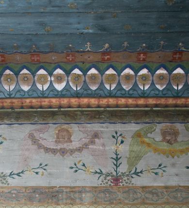 Zdjęcie nr 15: Ściany kościoła pokrywa dekoracja dywanowa, której w partiach stropu towarzyszą przedstawienia figuralne. W prezbiterium wykonano wyraźnie zaakcentowany fryz podstropowy, który od fasety oddziela profilowany i ornamentalnie malowany gzyms. Fryz na baldoniebieskim tle pokryto naprzemiennie rozmieszczonymi uskrzydlonymi główkami aniołów z niskimi wazonami z rozłożonymi trzema gałązkami kwitnących lilii. Poniżej barwnej bordiury oddzielającej fryz od pozostałych partii ścian, wykonano zgeometryzowaną dekorację utworzoną z powtarzających się wielobocznych pól, w których znalazł się stylizowany motyw tego samego kwiatu. Fasetę w prezbiterium pokryto naprzemiennie, rytmicznie rozmieszczonymi stylizowanymi kwiatami na łodydze i bez łodygi, które obwiedziono migdałowymi otokami tworzą ornamentalny pas zamknięty u góry falującą, wąską bordiurą. Dekorację bladoniebieskiego stropu prezbiterium tworzy poprowadzona przy krawędziach srebrno-niebieska arkatura. W centrum na niebieskim tle usianym złotymi gwiazdkami wyobrażono zasiadającą na obłokach Trójcę Świętą obramienioną pofalowaną, wielobarwną bordiurą otoczoną stylizowaną, srebrną wicią akantową. Dekoracyjną wić przedzielono parą uskrzydlonych główek anielskich oraz czterema stylizowanymi pąkami fantazyjnych kwiatów. Frontalnie ujęty Bóg Ojciec został wyobrażony jako siwobrody starzec o bujnych siwych włosach. W prawej ręce trzyma złote berło, lewą zaś wyciąga przed siebie. Bóg Ojciec prawą nogę opiera o niebieski glob, natomiast lewą, wyprostowaną wspiera się o obłoki. Jego twarz charakteryzuje jasna karnacja, szeroko otwarte oczy, wydatny nos i usta. Wokół głowy widać trójkąt. Obok Boga Ojca z lewej strony siedzi Chrystus, który obie nogi wspiera na globie. Zbawiciel trzymający oburącz na wysokości ramienia drewniany krzyż został przedstawiony w trzech czwartych w prawo. Jest to dojrzały mężczyzna o pociągłej twarzy okolonej brązowymi, długimi włosami spływającymi na ramiona oraz brodą. Ma jasną karnację, prosty nos i wydatne usta. Jego melancholijne spojrzenie jest skierowane w bok. Nagi tors okrywa bladoszkarłatny płaszcz odsłaniający prawą nogę. Na dłoniach i stopach widoczne są rany po gwoździach. Głowę Jezusa okala świetlisty nimb. Powyżej obu postaci unosi się gołębica Ducha Świętego w promienistym otoku. Niebieski glob na tle obłoków unoszą dwa aniołki w przepaskach. W nawie kościoła dekorację tworzą trzy różnej szerokości poziome pasy ornamentalne ułożone ponad jednolitą, szarą lamperią. Najszerszy pas pokryto na jasnym tle zgeometryzowanym ornamentem utworzonym z ciemnocynobrowych, migdałowo kształtowanych pól naśladujących wić roślinną. Powyżej rozmieszczono rytmicznie ułożone stylizowane krzyże równoramienne w fantazyjnych, wielobarwnych obramieniach. Najwyższy pas, odpowiadający fryzowi podstropowemu w prezbiterium pokryto wicią winnej latorośli z wplecionymi pękami kłosów. Tą część dekoracji zamyka profilowany gzyms ozdobiony wielobarwnym ornamentem zgeomertyzowanym. Rozpościerającą się ponad nim fasetę ozdobiono rytmicznie rozmieszczonym białym kwiatem na łodydze naśladujący w kształcie grzebień wodny. Dekorację bladoniebieskiego stropu artykułowano za pomocą srebrnej arkatury poprowadzonej przy jego krawędziach oraz ornamentalnych pasów utworzonych z motywów floralnych i geometrycznych, tworzących czworoboczną ramę dla centralnej kompozycji figuralnej. W narożach tego obramienia umieszczono medaliony ukazujące w popiersiach czterech ewangelistów malowanych na blasze miedzianej. Medaliony otaczają stylizowane gałązki dębowe z żołędziami. Każdego ewangelistę identyfikuje jego atrybut. W centrum stropu na bladozielonym tle usianym srebrnymi gwiazdkami namalowano wydłużone owalne obramienie utworzone z szerokiego wieńca różanego o bardzo drobnych kwiatach różanych i listkach. W jego centrum na tle bladoniebieskich obłoków ze złotymi gwiazdkami, bezpośrednio na deskach stropu, przedstawiono stojącą Madonnę z Dzieciątkiem, która depcze węża. Dodatkowo postać Marii zdaje się stać na rozpiętym różańcu. Maria ujęta frontalnie w kontrapoście obiema rękami podtrzymuje na prawym ramieniu Dzieciątko. Ma pociągłą twarz o jasnej karnacji, prostym nosie i wąskich ustach. Spojrzenie jej przymkniętych oczu jest skierowane w dół. Na głowie ma chustę szczelnie okrywającą jej włosy oraz złotą koronę o niskich sterczynach. Madonna ubrana jest w bladoczerwoną suknię odsłaniającą bosą stopę, którą depcze węża. Spod sukni na piersi widać białą koszulę. Na ramionach ma błękitny płaszcz podbity jasną ochrą, spięty na piersi podłużną, złotą klamrą. Trzymane przez nią Dzieciątko ma rozłożone ręce z uniesionymi dłońmi. Jego głowa ujęta jest w trzech czwartych w prawo. Otaczają ją jasno brązowe, krótkie włosy. Mały Jezus ubrany jest w jasną tunikę odsłaniającą jego bose stopy. Wokół Matki Boskiej rozmieszczono owalne medaliony z blachy miedzianej malowane w piętnaście tajemnic Różańca świętego. Ich narracja rozpoczyna się nad głową Madonny. Każda ze scen oparta jest o kilkuosobową kompozycję. Idąc w lewo namalowano: Zwiastowanie Marii, Nawiedzenie św. Elżbiety, Boże Narodzenie, Ofiarowanie w świątyni, Znalezienie w świątyni, Modlitwa w Ogrójcu, Biczowanie, Cierniem ukoronowanie, Niesienie krzyża, Ukrzyżowanie, Zmartwychwstanie, Wniebowstąpienie, Zesłanie Ducha Świętego, Wniebowzięcie Marii, Ukoronowanie Marii w niebie. Pomiędzy medalionami namalowano białą i czerwoną różę. Jasny parapet chóru muzycznego udekorowano naprzemiennie rozmieszczonymi rytmicznie stylizowanymi łodygami zakończonymi białymi pąkami i krzyżami równoramiennymi wpisanymi w koło. W aneksie południowym namalowano na ścianie kazanie św. Franciszka z Asyżu do ptaków. Kompozycja uzyskała kształt wydłużonego, stojącego prostokąta zamkniętego półkoliście i obwiedzionego falującą bordiurą z motywem łączących się krzyżyków. W jej centrum wyobrażono siedzącego na skalnym podłożu św. Franciszka ujętego w trzech czwartych w lewo. Święty prawą rękę unosi w geście mowy lewą zaś swobodnie trzyma na kolanach. Jego widoczną z profilu twarz o jasnej karnacji i prostym nosie okala siwiejąca broda i włosy z wyciętą tonsurą. Ubrany jest w brązowy habit przewiązany sznurem. Przed Franciszkiem przedstawiono stadko słuchających świetego ptaków. W prawym rogu kompozycji namalowano drewniane taczki. Z tyłu za świętym na tle skał rośnie drzewo. Inne drzewo wyobrażono z lewej strony, nieco z tyłu, na tle nieba pokrytego baldoniebieskimi obłokami. U góry z lewej strony na tle obłoków widać niewielką postać Madonny z Dzieciątkiem w typie Matki Boskiej Częstochowskiej.      