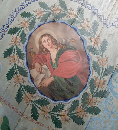 Zdjęcie nr 18: Ściany kościoła pokrywa dekoracja dywanowa, której w partiach stropu towarzyszą przedstawienia figuralne. W prezbiterium wykonano wyraźnie zaakcentowany fryz podstropowy, który od fasety oddziela profilowany i ornamentalnie malowany gzyms. Fryz na baldoniebieskim tle pokryto naprzemiennie rozmieszczonymi uskrzydlonymi główkami aniołów z niskimi wazonami z rozłożonymi trzema gałązkami kwitnących lilii. Poniżej barwnej bordiury oddzielającej fryz od pozostałych partii ścian, wykonano zgeometryzowaną dekorację utworzoną z powtarzających się wielobocznych pól, w których znalazł się stylizowany motyw tego samego kwiatu. Fasetę w prezbiterium pokryto naprzemiennie, rytmicznie rozmieszczonymi stylizowanymi kwiatami na łodydze i bez łodygi, które obwiedziono migdałowymi otokami tworzą ornamentalny pas zamknięty u góry falującą, wąską bordiurą. Dekorację bladoniebieskiego stropu prezbiterium tworzy poprowadzona przy krawędziach srebrno-niebieska arkatura. W centrum na niebieskim tle usianym złotymi gwiazdkami wyobrażono zasiadającą na obłokach Trójcę Świętą obramienioną pofalowaną, wielobarwną bordiurą otoczoną stylizowaną, srebrną wicią akantową. Dekoracyjną wić przedzielono parą uskrzydlonych główek anielskich oraz czterema stylizowanymi pąkami fantazyjnych kwiatów. Frontalnie ujęty Bóg Ojciec został wyobrażony jako siwobrody starzec o bujnych siwych włosach. W prawej ręce trzyma złote berło, lewą zaś wyciąga przed siebie. Bóg Ojciec prawą nogę opiera o niebieski glob, natomiast lewą, wyprostowaną wspiera się o obłoki. Jego twarz charakteryzuje jasna karnacja, szeroko otwarte oczy, wydatny nos i usta. Wokół głowy widać trójkąt. Obok Boga Ojca z lewej strony siedzi Chrystus, który obie nogi wspiera na globie. Zbawiciel trzymający oburącz na wysokości ramienia drewniany krzyż został przedstawiony w trzech czwartych w prawo. Jest to dojrzały mężczyzna o pociągłej twarzy okolonej brązowymi, długimi włosami spływającymi na ramiona oraz brodą. Ma jasną karnację, prosty nos i wydatne usta. Jego melancholijne spojrzenie jest skierowane w bok. Nagi tors okrywa bladoszkarłatny płaszcz odsłaniający prawą nogę. Na dłoniach i stopach widoczne są rany po gwoździach. Głowę Jezusa okala świetlisty nimb. Powyżej obu postaci unosi się gołębica Ducha Świętego w promienistym otoku. Niebieski glob na tle obłoków unoszą dwa aniołki w przepaskach. W nawie kościoła dekorację tworzą trzy różnej szerokości poziome pasy ornamentalne ułożone ponad jednolitą, szarą lamperią. Najszerszy pas pokryto na jasnym tle zgeometryzowanym ornamentem utworzonym z ciemnocynobrowych, migdałowo kształtowanych pól naśladujących wić roślinną. Powyżej rozmieszczono rytmicznie ułożone stylizowane krzyże równoramienne w fantazyjnych, wielobarwnych obramieniach. Najwyższy pas, odpowiadający fryzowi podstropowemu w prezbiterium pokryto wicią winnej latorośli z wplecionymi pękami kłosów. Tą część dekoracji zamyka profilowany gzyms ozdobiony wielobarwnym ornamentem zgeomertyzowanym. Rozpościerającą się ponad nim fasetę ozdobiono rytmicznie rozmieszczonym białym kwiatem na łodydze naśladujący w kształcie grzebień wodny. Dekorację bladoniebieskiego stropu artykułowano za pomocą srebrnej arkatury poprowadzonej przy jego krawędziach oraz ornamentalnych pasów utworzonych z motywów floralnych i geometrycznych, tworzących czworoboczną ramę dla centralnej kompozycji figuralnej. W narożach tego obramienia umieszczono medaliony ukazujące w popiersiach czterech ewangelistów malowanych na blasze miedzianej. Medaliony otaczają stylizowane gałązki dębowe z żołędziami. Każdego ewangelistę identyfikuje jego atrybut. W centrum stropu na bladozielonym tle usianym srebrnymi gwiazdkami namalowano wydłużone owalne obramienie utworzone z szerokiego wieńca różanego o bardzo drobnych kwiatach różanych i listkach. W jego centrum na tle bladoniebieskich obłoków ze złotymi gwiazdkami, bezpośrednio na deskach stropu, przedstawiono stojącą Madonnę z Dzieciątkiem, która depcze węża. Dodatkowo postać Marii zdaje się stać na rozpiętym różańcu. Maria ujęta frontalnie w kontrapoście obiema rękami podtrzymuje na prawym ramieniu Dzieciątko. Ma pociągłą twarz o jasnej karnacji, prostym nosie i wąskich ustach. Spojrzenie jej przymkniętych oczu jest skierowane w dół. Na głowie ma chustę szczelnie okrywającą jej włosy oraz złotą koronę o niskich sterczynach. Madonna ubrana jest w bladoczerwoną suknię odsłaniającą bosą stopę, którą depcze węża. Spod sukni na piersi widać białą koszulę. Na ramionach ma błękitny płaszcz podbity jasną ochrą, spięty na piersi podłużną, złotą klamrą. Trzymane przez nią Dzieciątko ma rozłożone ręce z uniesionymi dłońmi. Jego głowa ujęta jest w trzech czwartych w prawo. Otaczają ją jasno brązowe, krótkie włosy. Mały Jezus ubrany jest w jasną tunikę odsłaniającą jego bose stopy. Wokół Matki Boskiej rozmieszczono owalne medaliony z blachy miedzianej malowane w piętnaście tajemnic Różańca świętego. Ich narracja rozpoczyna się nad głową Madonny. Każda ze scen oparta jest o kilkuosobową kompozycję. Idąc w lewo namalowano: Zwiastowanie Marii, Nawiedzenie św. Elżbiety, Boże Narodzenie, Ofiarowanie w świątyni, Znalezienie w świątyni, Modlitwa w Ogrójcu, Biczowanie, Cierniem ukoronowanie, Niesienie krzyża, Ukrzyżowanie, Zmartwychwstanie, Wniebowstąpienie, Zesłanie Ducha Świętego, Wniebowzięcie Marii, Ukoronowanie Marii w niebie. Pomiędzy medalionami namalowano białą i czerwoną różę. Jasny parapet chóru muzycznego udekorowano naprzemiennie rozmieszczonymi rytmicznie stylizowanymi łodygami zakończonymi białymi pąkami i krzyżami równoramiennymi wpisanymi w koło. W aneksie południowym namalowano na ścianie kazanie św. Franciszka z Asyżu do ptaków. Kompozycja uzyskała kształt wydłużonego, stojącego prostokąta zamkniętego półkoliście i obwiedzionego falującą bordiurą z motywem łączących się krzyżyków. W jej centrum wyobrażono siedzącego na skalnym podłożu św. Franciszka ujętego w trzech czwartych w lewo. Święty prawą rękę unosi w geście mowy lewą zaś swobodnie trzyma na kolanach. Jego widoczną z profilu twarz o jasnej karnacji i prostym nosie okala siwiejąca broda i włosy z wyciętą tonsurą. Ubrany jest w brązowy habit przewiązany sznurem. Przed Franciszkiem przedstawiono stadko słuchających świetego ptaków. W prawym rogu kompozycji namalowano drewniane taczki. Z tyłu za świętym na tle skał rośnie drzewo. Inne drzewo wyobrażono z lewej strony, nieco z tyłu, na tle nieba pokrytego baldoniebieskimi obłokami. U góry z lewej strony na tle obłoków widać niewielką postać Madonny z Dzieciątkiem w typie Matki Boskiej Częstochowskiej.      