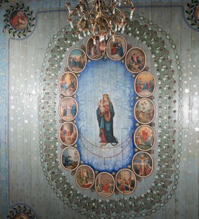 Zdjęcie nr 13: Ściany kościoła pokrywa dekoracja dywanowa, której w partiach stropu towarzyszą przedstawienia figuralne. W prezbiterium wykonano wyraźnie zaakcentowany fryz podstropowy, który od fasety oddziela profilowany i ornamentalnie malowany gzyms. Fryz na baldoniebieskim tle pokryto naprzemiennie rozmieszczonymi uskrzydlonymi główkami aniołów z niskimi wazonami z rozłożonymi trzema gałązkami kwitnących lilii. Poniżej barwnej bordiury oddzielającej fryz od pozostałych partii ścian, wykonano zgeometryzowaną dekorację utworzoną z powtarzających się wielobocznych pól, w których znalazł się stylizowany motyw tego samego kwiatu. Fasetę w prezbiterium pokryto naprzemiennie, rytmicznie rozmieszczonymi stylizowanymi kwiatami na łodydze i bez łodygi, które obwiedziono migdałowymi otokami tworzą ornamentalny pas zamknięty u góry falującą, wąską bordiurą. Dekorację bladoniebieskiego stropu prezbiterium tworzy poprowadzona przy krawędziach srebrno-niebieska arkatura. W centrum na niebieskim tle usianym złotymi gwiazdkami wyobrażono zasiadającą na obłokach Trójcę Świętą obramienioną pofalowaną, wielobarwną bordiurą otoczoną stylizowaną, srebrną wicią akantową. Dekoracyjną wić przedzielono parą uskrzydlonych główek anielskich oraz czterema stylizowanymi pąkami fantazyjnych kwiatów. Frontalnie ujęty Bóg Ojciec został wyobrażony jako siwobrody starzec o bujnych siwych włosach. W prawej ręce trzyma złote berło, lewą zaś wyciąga przed siebie. Bóg Ojciec prawą nogę opiera o niebieski glob, natomiast lewą, wyprostowaną wspiera się o obłoki. Jego twarz charakteryzuje jasna karnacja, szeroko otwarte oczy, wydatny nos i usta. Wokół głowy widać trójkąt. Obok Boga Ojca z lewej strony siedzi Chrystus, który obie nogi wspiera na globie. Zbawiciel trzymający oburącz na wysokości ramienia drewniany krzyż został przedstawiony w trzech czwartych w prawo. Jest to dojrzały mężczyzna o pociągłej twarzy okolonej brązowymi, długimi włosami spływającymi na ramiona oraz brodą. Ma jasną karnację, prosty nos i wydatne usta. Jego melancholijne spojrzenie jest skierowane w bok. Nagi tors okrywa bladoszkarłatny płaszcz odsłaniający prawą nogę. Na dłoniach i stopach widoczne są rany po gwoździach. Głowę Jezusa okala świetlisty nimb. Powyżej obu postaci unosi się gołębica Ducha Świętego w promienistym otoku. Niebieski glob na tle obłoków unoszą dwa aniołki w przepaskach. W nawie kościoła dekorację tworzą trzy różnej szerokości poziome pasy ornamentalne ułożone ponad jednolitą, szarą lamperią. Najszerszy pas pokryto na jasnym tle zgeometryzowanym ornamentem utworzonym z ciemnocynobrowych, migdałowo kształtowanych pól naśladujących wić roślinną. Powyżej rozmieszczono rytmicznie ułożone stylizowane krzyże równoramienne w fantazyjnych, wielobarwnych obramieniach. Najwyższy pas, odpowiadający fryzowi podstropowemu w prezbiterium pokryto wicią winnej latorośli z wplecionymi pękami kłosów. Tą część dekoracji zamyka profilowany gzyms ozdobiony wielobarwnym ornamentem zgeomertyzowanym. Rozpościerającą się ponad nim fasetę ozdobiono rytmicznie rozmieszczonym białym kwiatem na łodydze naśladujący w kształcie grzebień wodny. Dekorację bladoniebieskiego stropu artykułowano za pomocą srebrnej arkatury poprowadzonej przy jego krawędziach oraz ornamentalnych pasów utworzonych z motywów floralnych i geometrycznych, tworzących czworoboczną ramę dla centralnej kompozycji figuralnej. W narożach tego obramienia umieszczono medaliony ukazujące w popiersiach czterech ewangelistów malowanych na blasze miedzianej. Medaliony otaczają stylizowane gałązki dębowe z żołędziami. Każdego ewangelistę identyfikuje jego atrybut. W centrum stropu na bladozielonym tle usianym srebrnymi gwiazdkami namalowano wydłużone owalne obramienie utworzone z szerokiego wieńca różanego o bardzo drobnych kwiatach różanych i listkach. W jego centrum na tle bladoniebieskich obłoków ze złotymi gwiazdkami, bezpośrednio na deskach stropu, przedstawiono stojącą Madonnę z Dzieciątkiem, która depcze węża. Dodatkowo postać Marii zdaje się stać na rozpiętym różańcu. Maria ujęta frontalnie w kontrapoście obiema rękami podtrzymuje na prawym ramieniu Dzieciątko. Ma pociągłą twarz o jasnej karnacji, prostym nosie i wąskich ustach. Spojrzenie jej przymkniętych oczu jest skierowane w dół. Na głowie ma chustę szczelnie okrywającą jej włosy oraz złotą koronę o niskich sterczynach. Madonna ubrana jest w bladoczerwoną suknię odsłaniającą bosą stopę, którą depcze węża. Spod sukni na piersi widać białą koszulę. Na ramionach ma błękitny płaszcz podbity jasną ochrą, spięty na piersi podłużną, złotą klamrą. Trzymane przez nią Dzieciątko ma rozłożone ręce z uniesionymi dłońmi. Jego głowa ujęta jest w trzech czwartych w prawo. Otaczają ją jasno brązowe, krótkie włosy. Mały Jezus ubrany jest w jasną tunikę odsłaniającą jego bose stopy. Wokół Matki Boskiej rozmieszczono owalne medaliony z blachy miedzianej malowane w piętnaście tajemnic Różańca świętego. Ich narracja rozpoczyna się nad głową Madonny. Każda ze scen oparta jest o kilkuosobową kompozycję. Idąc w lewo namalowano: Zwiastowanie Marii, Nawiedzenie św. Elżbiety, Boże Narodzenie, Ofiarowanie w świątyni, Znalezienie w świątyni, Modlitwa w Ogrójcu, Biczowanie, Cierniem ukoronowanie, Niesienie krzyża, Ukrzyżowanie, Zmartwychwstanie, Wniebowstąpienie, Zesłanie Ducha Świętego, Wniebowzięcie Marii, Ukoronowanie Marii w niebie. Pomiędzy medalionami namalowano białą i czerwoną różę. Jasny parapet chóru muzycznego udekorowano naprzemiennie rozmieszczonymi rytmicznie stylizowanymi łodygami zakończonymi białymi pąkami i krzyżami równoramiennymi wpisanymi w koło. W aneksie południowym namalowano na ścianie kazanie św. Franciszka z Asyżu do ptaków. Kompozycja uzyskała kształt wydłużonego, stojącego prostokąta zamkniętego półkoliście i obwiedzionego falującą bordiurą z motywem łączących się krzyżyków. W jej centrum wyobrażono siedzącego na skalnym podłożu św. Franciszka ujętego w trzech czwartych w lewo. Święty prawą rękę unosi w geście mowy lewą zaś swobodnie trzyma na kolanach. Jego widoczną z profilu twarz o jasnej karnacji i prostym nosie okala siwiejąca broda i włosy z wyciętą tonsurą. Ubrany jest w brązowy habit przewiązany sznurem. Przed Franciszkiem przedstawiono stadko słuchających świetego ptaków. W prawym rogu kompozycji namalowano drewniane taczki. Z tyłu za świętym na tle skał rośnie drzewo. Inne drzewo wyobrażono z lewej strony, nieco z tyłu, na tle nieba pokrytego baldoniebieskimi obłokami. U góry z lewej strony na tle obłoków widać niewielką postać Madonny z Dzieciątkiem w typie Matki Boskiej Częstochowskiej.      