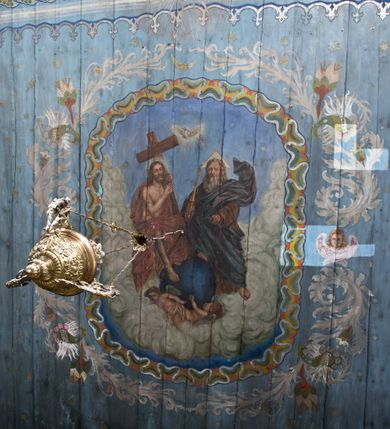 Zdjęcie nr 16: Ściany kościoła pokrywa dekoracja dywanowa, której w partiach stropu towarzyszą przedstawienia figuralne. W prezbiterium wykonano wyraźnie zaakcentowany fryz podstropowy, który od fasety oddziela profilowany i ornamentalnie malowany gzyms. Fryz na baldoniebieskim tle pokryto naprzemiennie rozmieszczonymi uskrzydlonymi główkami aniołów z niskimi wazonami z rozłożonymi trzema gałązkami kwitnących lilii. Poniżej barwnej bordiury oddzielającej fryz od pozostałych partii ścian, wykonano zgeometryzowaną dekorację utworzoną z powtarzających się wielobocznych pól, w których znalazł się stylizowany motyw tego samego kwiatu. Fasetę w prezbiterium pokryto naprzemiennie, rytmicznie rozmieszczonymi stylizowanymi kwiatami na łodydze i bez łodygi, które obwiedziono migdałowymi otokami tworzą ornamentalny pas zamknięty u góry falującą, wąską bordiurą. Dekorację bladoniebieskiego stropu prezbiterium tworzy poprowadzona przy krawędziach srebrno-niebieska arkatura. W centrum na niebieskim tle usianym złotymi gwiazdkami wyobrażono zasiadającą na obłokach Trójcę Świętą obramienioną pofalowaną, wielobarwną bordiurą otoczoną stylizowaną, srebrną wicią akantową. Dekoracyjną wić przedzielono parą uskrzydlonych główek anielskich oraz czterema stylizowanymi pąkami fantazyjnych kwiatów. Frontalnie ujęty Bóg Ojciec został wyobrażony jako siwobrody starzec o bujnych siwych włosach. W prawej ręce trzyma złote berło, lewą zaś wyciąga przed siebie. Bóg Ojciec prawą nogę opiera o niebieski glob, natomiast lewą, wyprostowaną wspiera się o obłoki. Jego twarz charakteryzuje jasna karnacja, szeroko otwarte oczy, wydatny nos i usta. Wokół głowy widać trójkąt. Obok Boga Ojca z lewej strony siedzi Chrystus, który obie nogi wspiera na globie. Zbawiciel trzymający oburącz na wysokości ramienia drewniany krzyż został przedstawiony w trzech czwartych w prawo. Jest to dojrzały mężczyzna o pociągłej twarzy okolonej brązowymi, długimi włosami spływającymi na ramiona oraz brodą. Ma jasną karnację, prosty nos i wydatne usta. Jego melancholijne spojrzenie jest skierowane w bok. Nagi tors okrywa bladoszkarłatny płaszcz odsłaniający prawą nogę. Na dłoniach i stopach widoczne są rany po gwoździach. Głowę Jezusa okala świetlisty nimb. Powyżej obu postaci unosi się gołębica Ducha Świętego w promienistym otoku. Niebieski glob na tle obłoków unoszą dwa aniołki w przepaskach. W nawie kościoła dekorację tworzą trzy różnej szerokości poziome pasy ornamentalne ułożone ponad jednolitą, szarą lamperią. Najszerszy pas pokryto na jasnym tle zgeometryzowanym ornamentem utworzonym z ciemnocynobrowych, migdałowo kształtowanych pól naśladujących wić roślinną. Powyżej rozmieszczono rytmicznie ułożone stylizowane krzyże równoramienne w fantazyjnych, wielobarwnych obramieniach. Najwyższy pas, odpowiadający fryzowi podstropowemu w prezbiterium pokryto wicią winnej latorośli z wplecionymi pękami kłosów. Tą część dekoracji zamyka profilowany gzyms ozdobiony wielobarwnym ornamentem zgeomertyzowanym. Rozpościerającą się ponad nim fasetę ozdobiono rytmicznie rozmieszczonym białym kwiatem na łodydze naśladujący w kształcie grzebień wodny. Dekorację bladoniebieskiego stropu artykułowano za pomocą srebrnej arkatury poprowadzonej przy jego krawędziach oraz ornamentalnych pasów utworzonych z motywów floralnych i geometrycznych, tworzących czworoboczną ramę dla centralnej kompozycji figuralnej. W narożach tego obramienia umieszczono medaliony ukazujące w popiersiach czterech ewangelistów malowanych na blasze miedzianej. Medaliony otaczają stylizowane gałązki dębowe z żołędziami. Każdego ewangelistę identyfikuje jego atrybut. W centrum stropu na bladozielonym tle usianym srebrnymi gwiazdkami namalowano wydłużone owalne obramienie utworzone z szerokiego wieńca różanego o bardzo drobnych kwiatach różanych i listkach. W jego centrum na tle bladoniebieskich obłoków ze złotymi gwiazdkami, bezpośrednio na deskach stropu, przedstawiono stojącą Madonnę z Dzieciątkiem, która depcze węża. Dodatkowo postać Marii zdaje się stać na rozpiętym różańcu. Maria ujęta frontalnie w kontrapoście obiema rękami podtrzymuje na prawym ramieniu Dzieciątko. Ma pociągłą twarz o jasnej karnacji, prostym nosie i wąskich ustach. Spojrzenie jej przymkniętych oczu jest skierowane w dół. Na głowie ma chustę szczelnie okrywającą jej włosy oraz złotą koronę o niskich sterczynach. Madonna ubrana jest w bladoczerwoną suknię odsłaniającą bosą stopę, którą depcze węża. Spod sukni na piersi widać białą koszulę. Na ramionach ma błękitny płaszcz podbity jasną ochrą, spięty na piersi podłużną, złotą klamrą. Trzymane przez nią Dzieciątko ma rozłożone ręce z uniesionymi dłońmi. Jego głowa ujęta jest w trzech czwartych w prawo. Otaczają ją jasno brązowe, krótkie włosy. Mały Jezus ubrany jest w jasną tunikę odsłaniającą jego bose stopy. Wokół Matki Boskiej rozmieszczono owalne medaliony z blachy miedzianej malowane w piętnaście tajemnic Różańca świętego. Ich narracja rozpoczyna się nad głową Madonny. Każda ze scen oparta jest o kilkuosobową kompozycję. Idąc w lewo namalowano: Zwiastowanie Marii, Nawiedzenie św. Elżbiety, Boże Narodzenie, Ofiarowanie w świątyni, Znalezienie w świątyni, Modlitwa w Ogrójcu, Biczowanie, Cierniem ukoronowanie, Niesienie krzyża, Ukrzyżowanie, Zmartwychwstanie, Wniebowstąpienie, Zesłanie Ducha Świętego, Wniebowzięcie Marii, Ukoronowanie Marii w niebie. Pomiędzy medalionami namalowano białą i czerwoną różę. Jasny parapet chóru muzycznego udekorowano naprzemiennie rozmieszczonymi rytmicznie stylizowanymi łodygami zakończonymi białymi pąkami i krzyżami równoramiennymi wpisanymi w koło. W aneksie południowym namalowano na ścianie kazanie św. Franciszka z Asyżu do ptaków. Kompozycja uzyskała kształt wydłużonego, stojącego prostokąta zamkniętego półkoliście i obwiedzionego falującą bordiurą z motywem łączących się krzyżyków. W jej centrum wyobrażono siedzącego na skalnym podłożu św. Franciszka ujętego w trzech czwartych w lewo. Święty prawą rękę unosi w geście mowy lewą zaś swobodnie trzyma na kolanach. Jego widoczną z profilu twarz o jasnej karnacji i prostym nosie okala siwiejąca broda i włosy z wyciętą tonsurą. Ubrany jest w brązowy habit przewiązany sznurem. Przed Franciszkiem przedstawiono stadko słuchających świetego ptaków. W prawym rogu kompozycji namalowano drewniane taczki. Z tyłu za świętym na tle skał rośnie drzewo. Inne drzewo wyobrażono z lewej strony, nieco z tyłu, na tle nieba pokrytego baldoniebieskimi obłokami. U góry z lewej strony na tle obłoków widać niewielką postać Madonny z Dzieciątkiem w typie Matki Boskiej Częstochowskiej.      