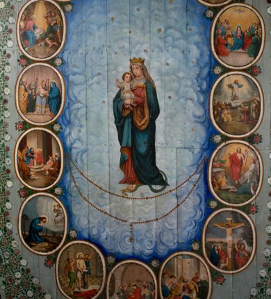 Zdjęcie nr 1: Ściany kościoła pokrywa dekoracja dywanowa, której w partiach stropu towarzyszą przedstawienia figuralne. W prezbiterium wykonano wyraźnie zaakcentowany fryz podstropowy, który od fasety oddziela profilowany i ornamentalnie malowany gzyms. Fryz na baldoniebieskim tle pokryto naprzemiennie rozmieszczonymi uskrzydlonymi główkami aniołów z niskimi wazonami z rozłożonymi trzema gałązkami kwitnących lilii. Poniżej barwnej bordiury oddzielającej fryz od pozostałych partii ścian, wykonano zgeometryzowaną dekorację utworzoną z powtarzających się wielobocznych pól, w których znalazł się stylizowany motyw tego samego kwiatu. Fasetę w prezbiterium pokryto naprzemiennie, rytmicznie rozmieszczonymi stylizowanymi kwiatami na łodydze i bez łodygi, które obwiedziono migdałowymi otokami tworzą ornamentalny pas zamknięty u góry falującą, wąską bordiurą. Dekorację bladoniebieskiego stropu prezbiterium tworzy poprowadzona przy krawędziach srebrno-niebieska arkatura. W centrum na niebieskim tle usianym złotymi gwiazdkami wyobrażono zasiadającą na obłokach Trójcę Świętą obramienioną pofalowaną, wielobarwną bordiurą otoczoną stylizowaną, srebrną wicią akantową. Dekoracyjną wić przedzielono parą uskrzydlonych główek anielskich oraz czterema stylizowanymi pąkami fantazyjnych kwiatów. Frontalnie ujęty Bóg Ojciec został wyobrażony jako siwobrody starzec o bujnych siwych włosach. W prawej ręce trzyma złote berło, lewą zaś wyciąga przed siebie. Bóg Ojciec prawą nogę opiera o niebieski glob, natomiast lewą, wyprostowaną wspiera się o obłoki. Jego twarz charakteryzuje jasna karnacja, szeroko otwarte oczy, wydatny nos i usta. Wokół głowy widać trójkąt. Obok Boga Ojca z lewej strony siedzi Chrystus, który obie nogi wspiera na globie. Zbawiciel trzymający oburącz na wysokości ramienia drewniany krzyż został przedstawiony w trzech czwartych w prawo. Jest to dojrzały mężczyzna o pociągłej twarzy okolonej brązowymi, długimi włosami spływającymi na ramiona oraz brodą. Ma jasną karnację, prosty nos i wydatne usta. Jego melancholijne spojrzenie jest skierowane w bok. Nagi tors okrywa bladoszkarłatny płaszcz odsłaniający prawą nogę. Na dłoniach i stopach widoczne są rany po gwoździach. Głowę Jezusa okala świetlisty nimb. Powyżej obu postaci unosi się gołębica Ducha Świętego w promienistym otoku. Niebieski glob na tle obłoków unoszą dwa aniołki w przepaskach. W nawie kościoła dekorację tworzą trzy różnej szerokości poziome pasy ornamentalne ułożone ponad jednolitą, szarą lamperią. Najszerszy pas pokryto na jasnym tle zgeometryzowanym ornamentem utworzonym z ciemnocynobrowych, migdałowo kształtowanych pól naśladujących wić roślinną. Powyżej rozmieszczono rytmicznie ułożone stylizowane krzyże równoramienne w fantazyjnych, wielobarwnych obramieniach. Najwyższy pas, odpowiadający fryzowi podstropowemu w prezbiterium pokryto wicią winnej latorośli z wplecionymi pękami kłosów. Tą część dekoracji zamyka profilowany gzyms ozdobiony wielobarwnym ornamentem zgeomertyzowanym. Rozpościerającą się ponad nim fasetę ozdobiono rytmicznie rozmieszczonym białym kwiatem na łodydze naśladujący w kształcie grzebień wodny. Dekorację bladoniebieskiego stropu artykułowano za pomocą srebrnej arkatury poprowadzonej przy jego krawędziach oraz ornamentalnych pasów utworzonych z motywów floralnych i geometrycznych, tworzących czworoboczną ramę dla centralnej kompozycji figuralnej. W narożach tego obramienia umieszczono medaliony ukazujące w popiersiach czterech ewangelistów malowanych na blasze miedzianej. Medaliony otaczają stylizowane gałązki dębowe z żołędziami. Każdego ewangelistę identyfikuje jego atrybut. W centrum stropu na bladozielonym tle usianym srebrnymi gwiazdkami namalowano wydłużone owalne obramienie utworzone z szerokiego wieńca różanego o bardzo drobnych kwiatach różanych i listkach. W jego centrum na tle bladoniebieskich obłoków ze złotymi gwiazdkami, bezpośrednio na deskach stropu, przedstawiono stojącą Madonnę z Dzieciątkiem, która depcze węża. Dodatkowo postać Marii zdaje się stać na rozpiętym różańcu. Maria ujęta frontalnie w kontrapoście obiema rękami podtrzymuje na prawym ramieniu Dzieciątko. Ma pociągłą twarz o jasnej karnacji, prostym nosie i wąskich ustach. Spojrzenie jej przymkniętych oczu jest skierowane w dół. Na głowie ma chustę szczelnie okrywającą jej włosy oraz złotą koronę o niskich sterczynach. Madonna ubrana jest w bladoczerwoną suknię odsłaniającą bosą stopę, którą depcze węża. Spod sukni na piersi widać białą koszulę. Na ramionach ma błękitny płaszcz podbity jasną ochrą, spięty na piersi podłużną, złotą klamrą. Trzymane przez nią Dzieciątko ma rozłożone ręce z uniesionymi dłońmi. Jego głowa ujęta jest w trzech czwartych w prawo. Otaczają ją jasno brązowe, krótkie włosy. Mały Jezus ubrany jest w jasną tunikę odsłaniającą jego bose stopy. Wokół Matki Boskiej rozmieszczono owalne medaliony z blachy miedzianej malowane w piętnaście tajemnic Różańca świętego. Ich narracja rozpoczyna się nad głową Madonny. Każda ze scen oparta jest o kilkuosobową kompozycję. Idąc w lewo namalowano: Zwiastowanie Marii, Nawiedzenie św. Elżbiety, Boże Narodzenie, Ofiarowanie w świątyni, Znalezienie w świątyni, Modlitwa w Ogrójcu, Biczowanie, Cierniem ukoronowanie, Niesienie krzyża, Ukrzyżowanie, Zmartwychwstanie, Wniebowstąpienie, Zesłanie Ducha Świętego, Wniebowzięcie Marii, Ukoronowanie Marii w niebie. Pomiędzy medalionami namalowano białą i czerwoną różę. Jasny parapet chóru muzycznego udekorowano naprzemiennie rozmieszczonymi rytmicznie stylizowanymi łodygami zakończonymi białymi pąkami i krzyżami równoramiennymi wpisanymi w koło. W aneksie południowym namalowano na ścianie kazanie św. Franciszka z Asyżu do ptaków. Kompozycja uzyskała kształt wydłużonego, stojącego prostokąta zamkniętego półkoliście i obwiedzionego falującą bordiurą z motywem łączących się krzyżyków. W jej centrum wyobrażono siedzącego na skalnym podłożu św. Franciszka ujętego w trzech czwartych w lewo. Święty prawą rękę unosi w geście mowy lewą zaś swobodnie trzyma na kolanach. Jego widoczną z profilu twarz o jasnej karnacji i prostym nosie okala siwiejąca broda i włosy z wyciętą tonsurą. Ubrany jest w brązowy habit przewiązany sznurem. Przed Franciszkiem przedstawiono stadko słuchających świetego ptaków. W prawym rogu kompozycji namalowano drewniane taczki. Z tyłu za świętym na tle skał rośnie drzewo. Inne drzewo wyobrażono z lewej strony, nieco z tyłu, na tle nieba pokrytego baldoniebieskimi obłokami. U góry z lewej strony na tle obłoków widać niewielką postać Madonny z Dzieciątkiem w typie Matki Boskiej Częstochowskiej.      