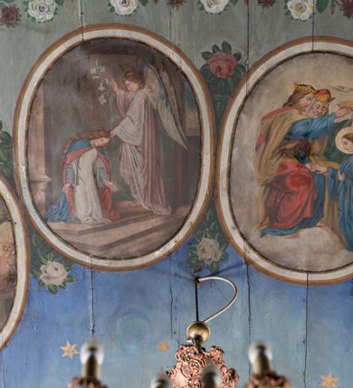 Zdjęcie nr 10: Ściany kościoła pokrywa dekoracja dywanowa, której w partiach stropu towarzyszą przedstawienia figuralne. W prezbiterium wykonano wyraźnie zaakcentowany fryz podstropowy, który od fasety oddziela profilowany i ornamentalnie malowany gzyms. Fryz na baldoniebieskim tle pokryto naprzemiennie rozmieszczonymi uskrzydlonymi główkami aniołów z niskimi wazonami z rozłożonymi trzema gałązkami kwitnących lilii. Poniżej barwnej bordiury oddzielającej fryz od pozostałych partii ścian, wykonano zgeometryzowaną dekorację utworzoną z powtarzających się wielobocznych pól, w których znalazł się stylizowany motyw tego samego kwiatu. Fasetę w prezbiterium pokryto naprzemiennie, rytmicznie rozmieszczonymi stylizowanymi kwiatami na łodydze i bez łodygi, które obwiedziono migdałowymi otokami tworzą ornamentalny pas zamknięty u góry falującą, wąską bordiurą. Dekorację bladoniebieskiego stropu prezbiterium tworzy poprowadzona przy krawędziach srebrno-niebieska arkatura. W centrum na niebieskim tle usianym złotymi gwiazdkami wyobrażono zasiadającą na obłokach Trójcę Świętą obramienioną pofalowaną, wielobarwną bordiurą otoczoną stylizowaną, srebrną wicią akantową. Dekoracyjną wić przedzielono parą uskrzydlonych główek anielskich oraz czterema stylizowanymi pąkami fantazyjnych kwiatów. Frontalnie ujęty Bóg Ojciec został wyobrażony jako siwobrody starzec o bujnych siwych włosach. W prawej ręce trzyma złote berło, lewą zaś wyciąga przed siebie. Bóg Ojciec prawą nogę opiera o niebieski glob, natomiast lewą, wyprostowaną wspiera się o obłoki. Jego twarz charakteryzuje jasna karnacja, szeroko otwarte oczy, wydatny nos i usta. Wokół głowy widać trójkąt. Obok Boga Ojca z lewej strony siedzi Chrystus, który obie nogi wspiera na globie. Zbawiciel trzymający oburącz na wysokości ramienia drewniany krzyż został przedstawiony w trzech czwartych w prawo. Jest to dojrzały mężczyzna o pociągłej twarzy okolonej brązowymi, długimi włosami spływającymi na ramiona oraz brodą. Ma jasną karnację, prosty nos i wydatne usta. Jego melancholijne spojrzenie jest skierowane w bok. Nagi tors okrywa bladoszkarłatny płaszcz odsłaniający prawą nogę. Na dłoniach i stopach widoczne są rany po gwoździach. Głowę Jezusa okala świetlisty nimb. Powyżej obu postaci unosi się gołębica Ducha Świętego w promienistym otoku. Niebieski glob na tle obłoków unoszą dwa aniołki w przepaskach. W nawie kościoła dekorację tworzą trzy różnej szerokości poziome pasy ornamentalne ułożone ponad jednolitą, szarą lamperią. Najszerszy pas pokryto na jasnym tle zgeometryzowanym ornamentem utworzonym z ciemnocynobrowych, migdałowo kształtowanych pól naśladujących wić roślinną. Powyżej rozmieszczono rytmicznie ułożone stylizowane krzyże równoramienne w fantazyjnych, wielobarwnych obramieniach. Najwyższy pas, odpowiadający fryzowi podstropowemu w prezbiterium pokryto wicią winnej latorośli z wplecionymi pękami kłosów. Tą część dekoracji zamyka profilowany gzyms ozdobiony wielobarwnym ornamentem zgeomertyzowanym. Rozpościerającą się ponad nim fasetę ozdobiono rytmicznie rozmieszczonym białym kwiatem na łodydze naśladujący w kształcie grzebień wodny. Dekorację bladoniebieskiego stropu artykułowano za pomocą srebrnej arkatury poprowadzonej przy jego krawędziach oraz ornamentalnych pasów utworzonych z motywów floralnych i geometrycznych, tworzących czworoboczną ramę dla centralnej kompozycji figuralnej. W narożach tego obramienia umieszczono medaliony ukazujące w popiersiach czterech ewangelistów malowanych na blasze miedzianej. Medaliony otaczają stylizowane gałązki dębowe z żołędziami. Każdego ewangelistę identyfikuje jego atrybut. W centrum stropu na bladozielonym tle usianym srebrnymi gwiazdkami namalowano wydłużone owalne obramienie utworzone z szerokiego wieńca różanego o bardzo drobnych kwiatach różanych i listkach. W jego centrum na tle bladoniebieskich obłoków ze złotymi gwiazdkami, bezpośrednio na deskach stropu, przedstawiono stojącą Madonnę z Dzieciątkiem, która depcze węża. Dodatkowo postać Marii zdaje się stać na rozpiętym różańcu. Maria ujęta frontalnie w kontrapoście obiema rękami podtrzymuje na prawym ramieniu Dzieciątko. Ma pociągłą twarz o jasnej karnacji, prostym nosie i wąskich ustach. Spojrzenie jej przymkniętych oczu jest skierowane w dół. Na głowie ma chustę szczelnie okrywającą jej włosy oraz złotą koronę o niskich sterczynach. Madonna ubrana jest w bladoczerwoną suknię odsłaniającą bosą stopę, którą depcze węża. Spod sukni na piersi widać białą koszulę. Na ramionach ma błękitny płaszcz podbity jasną ochrą, spięty na piersi podłużną, złotą klamrą. Trzymane przez nią Dzieciątko ma rozłożone ręce z uniesionymi dłońmi. Jego głowa ujęta jest w trzech czwartych w prawo. Otaczają ją jasno brązowe, krótkie włosy. Mały Jezus ubrany jest w jasną tunikę odsłaniającą jego bose stopy. Wokół Matki Boskiej rozmieszczono owalne medaliony z blachy miedzianej malowane w piętnaście tajemnic Różańca świętego. Ich narracja rozpoczyna się nad głową Madonny. Każda ze scen oparta jest o kilkuosobową kompozycję. Idąc w lewo namalowano: Zwiastowanie Marii, Nawiedzenie św. Elżbiety, Boże Narodzenie, Ofiarowanie w świątyni, Znalezienie w świątyni, Modlitwa w Ogrójcu, Biczowanie, Cierniem ukoronowanie, Niesienie krzyża, Ukrzyżowanie, Zmartwychwstanie, Wniebowstąpienie, Zesłanie Ducha Świętego, Wniebowzięcie Marii, Ukoronowanie Marii w niebie. Pomiędzy medalionami namalowano białą i czerwoną różę. Jasny parapet chóru muzycznego udekorowano naprzemiennie rozmieszczonymi rytmicznie stylizowanymi łodygami zakończonymi białymi pąkami i krzyżami równoramiennymi wpisanymi w koło. W aneksie południowym namalowano na ścianie kazanie św. Franciszka z Asyżu do ptaków. Kompozycja uzyskała kształt wydłużonego, stojącego prostokąta zamkniętego półkoliście i obwiedzionego falującą bordiurą z motywem łączących się krzyżyków. W jej centrum wyobrażono siedzącego na skalnym podłożu św. Franciszka ujętego w trzech czwartych w lewo. Święty prawą rękę unosi w geście mowy lewą zaś swobodnie trzyma na kolanach. Jego widoczną z profilu twarz o jasnej karnacji i prostym nosie okala siwiejąca broda i włosy z wyciętą tonsurą. Ubrany jest w brązowy habit przewiązany sznurem. Przed Franciszkiem przedstawiono stadko słuchających świetego ptaków. W prawym rogu kompozycji namalowano drewniane taczki. Z tyłu za świętym na tle skał rośnie drzewo. Inne drzewo wyobrażono z lewej strony, nieco z tyłu, na tle nieba pokrytego baldoniebieskimi obłokami. U góry z lewej strony na tle obłoków widać niewielką postać Madonny z Dzieciątkiem w typie Matki Boskiej Częstochowskiej.      
