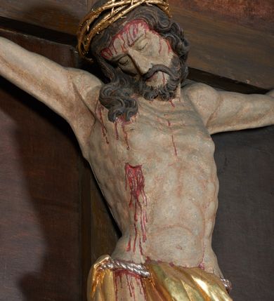 Zdjęcie nr 5: Krucyfiks w typie Christo morte; postać  Chrystusa umarłego przybitego do krzyża trzema gwoździami. Sylwetka w delikatnym zwisie, o silnie zaznaczonej anatomii ciała, z podkreśloną linią żeber i zaznaczonymi śladami męki. Perizonium w formie tkaniny przewiązanej na sznurze, sięgające kolan i odsłaniające prawe udo; z prawej strony spływające kaskadowo. Twarz Jezusa podłużna opadająca na prawe ramię, okolona zarostem; oczy zamknięte, nos podłużny, wystające kości policzkowe; włosy długie spływające na prawe ramię, na głowie złota korona cierniowa. Partie ciała polichromowane naturalistycznie, korona cierniowa i perizonium złocone.