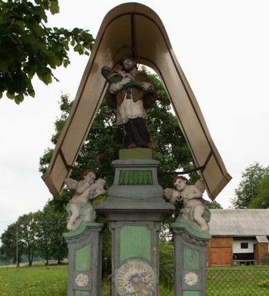 Zdjęcie nr 1: Kapliczka ustawiona na niskim prostopadłościennym podeście, dwukondygnacyjna. W dolnej partii trzy filary ustawione na masywnych cokołach, u dołu profilowane; boczne zdobione perełkowaniem, a u góry zamknięte falującym gzymsem i dekorowane kostkowaniem. Na ściankach frontowych płyciny w kształcie stojących prostokątów, na filarze środkowym tondo z obłokami i promienistą glorią, na filarach bocznych białe kwiaty. Na środkowym filarze, na wysokim profilowanym cokole, zdobionym z przodu prostokątną płyciną, malowaną w zielone liście, a u góry zamkniętym gzymsem dekorowanym kimationem ustawiona jest figura św. Jana Nepomucena. Święty ukazany w postawie stojącej w kontrapoście o esowato wygiętej sylwetce z krucyfiksem i palmą męczeństwa w dłoniach. Twarz szeroka z długim nosem, okolona krótką brodą. Święty ubrany jest czarną sutannę, białą rokietę i befkę oraz brązową almucję, na głowie ma założony czarny biret, a wokół głowy wieniec z pięciu gwiazd, na stopach bordowe buty. Na bocznych filarach ustawione są rzeźby dwóch aniołków, z których lewy trzyma kłódkę w dłoniach, a prawy klucz. Przed kapliczką leży płyta, silnie zniszczona z długą łacińską inskrypcją, słabo czytelną: „MAGNE […] ROGAS TIBI QVIS DICAT HAEC MOR[…] / […] / […] / […] HOC / [...] RES / […] CAV[…] / […] COLIT IN / […] MORE DIC[…]A / […]SOCER[…] / […]”. Kapliczka u góry nakryta metalowym, współczesnym daszkiem. 
