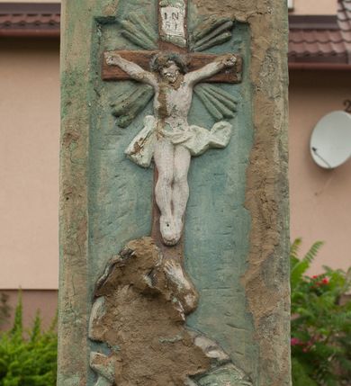 Zdjęcie nr 2: Kapliczka ustawiona na niskim prostopadłościennym postumencie w formie zdwojonego słupa, zamkniętego profilowanym gzymsem, na którego szczycie ustawiona jest figura Matki Boskiej z Dzieciątkiem. Na ściance frontowej cokołu wieniec z liści, w polu inskrypcja: „ANNO D[OMI]NI / 1759 DIE 1/ IUNI(?)”. Na cokole ustawiony czworoboczny filar zdobiony płaskorzeźbami. Na ściance frontowej w prostokątnej płycinie, zamkniętej łukiem nadwieszonym krucyfiks i św. Maria Magdalena, powyżej dwie uskrzydlone główki anielskie. Poniżej słaboczytelna inskrypcja: „(?)1815 DIE / 18. 8BRIS.” Na lewej ściance bocznej w analogicznej niszy rzeźba nierozpoznanej świętej, wyżej w tondzie uskrzydlona główka anielska. Na szczycie pod metalowym daszkiem zamocowanym na czterech prętach jest figura Matki Boskiej z Dzieciątkiem. Maria ukazana w postawie stojącej na globie ziemskim, prawą stopą depcząca węża, z Dzieciątkiem Jezus na prawym ręku. Twarz szeroka o sumarycznie oddanych rysach twarzy; włosy ciemnobrązowe, bujne, spływające na plecy. Ubrana jest w białą suknię oraz niebieski płaszcz, na stopach ma założone sandały. Dzieciątko zwrócone w prawo ze skrzyżowanymi nóżkami, rączkami chwytające matkę za szyję. Twarz o podobnych rysach, okolona krótkimi, jasnymi włosami. Ubrane jest w białą pieluszkę, owijającą biodra. Polichromia postaci w odsłoniętych partiach ciała naturalistyczna. 
