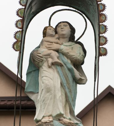 Zdjęcie nr 1: Kapliczka ustawiona na niskim prostopadłościennym postumencie w formie zdwojonego słupa, zamkniętego profilowanym gzymsem, na którego szczycie ustawiona jest figura Matki Boskiej z Dzieciątkiem. Na ściance frontowej cokołu wieniec z liści, w polu inskrypcja: „ANNO D[OMI]NI / 1759 DIE 1/ IUNI(?)”. Na cokole ustawiony czworoboczny filar zdobiony płaskorzeźbami. Na ściance frontowej w prostokątnej płycinie, zamkniętej łukiem nadwieszonym krucyfiks i św. Maria Magdalena, powyżej dwie uskrzydlone główki anielskie. Poniżej słaboczytelna inskrypcja: „(?)1815 DIE / 18. 8BRIS.” Na lewej ściance bocznej w analogicznej niszy rzeźba nierozpoznanej świętej, wyżej w tondzie uskrzydlona główka anielska. Na szczycie pod metalowym daszkiem zamocowanym na czterech prętach jest figura Matki Boskiej z Dzieciątkiem. Maria ukazana w postawie stojącej na globie ziemskim, prawą stopą depcząca węża, z Dzieciątkiem Jezus na prawym ręku. Twarz szeroka o sumarycznie oddanych rysach twarzy; włosy ciemnobrązowe, bujne, spływające na plecy. Ubrana jest w białą suknię oraz niebieski płaszcz, na stopach ma założone sandały. Dzieciątko zwrócone w prawo ze skrzyżowanymi nóżkami, rączkami chwytające matkę za szyję. Twarz o podobnych rysach, okolona krótkimi, jasnymi włosami. Ubrane jest w białą pieluszkę, owijającą biodra. Polichromia postaci w odsłoniętych partiach ciała naturalistyczna. 
