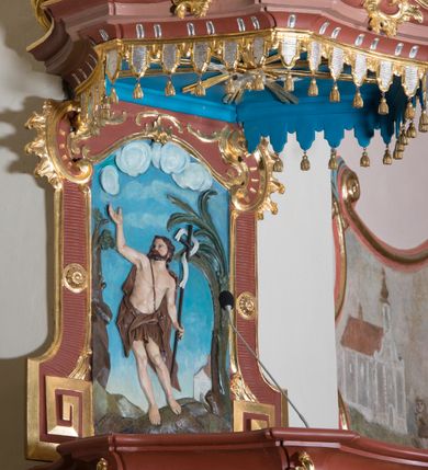 Zdjęcie nr 7: Ambona przyścienna, wisząca ustawiona przy północnej ścianie, dostępna jednobiegowymi schodami z balustradą od strony prezbiterium. Kosz na planie sześcioboku u dołu z ćwierćwałkiem, zdobionym na narożach dekoracją rokokową; u góry zamknięty profilowanym gzymsem. Ścianki artykułowane spływami wolutowymi o karbowanych trzonach. W polach prostokątne płyciny mieszczące płaskorzeźbione rocaille. Podwieszenie w formie stożka o wklęsło-wypukłych bokach, zakończonego płaskorzeźbioną rozetą. Ścianki flankowane w narożach analogicznie jak kosz, w polach trapezoidalne płyciny, przerwane pośrodku okręgiem z rozetką. Zaplecek prostokąty ujęty płaskim obramieniem o zgeometryzowanych wolutach w dolnych narożach oraz rocaille'm w górnych. Pośrodku płaskorzeźbione przedstawienie św. Jana Chrzciciela na tle pejzażu. Baldachim powtarzający plan kosza, zwieńczony wazonem ustawionym na sześciu spiętych kabłąkach. W podniebiu gołębica Ducha Świętego na tle promienistej glorii, na brzegach zawieszony lambrekin. Balustrada schodów pełna, od dołu ujęta półwałkiem, zamknięta profilowanym gzymsem, w polu dwie  płyciny w kształcie rombów, w których umieszczone są rocaille. Balustrada zakończona filarkiem, na którym ustawiony jest wazonik. Całość struktury ambony polichromowana w odcieniach brązu, płaskorzeźba wielobarwna, profilowania i detal architektoniczny pozłacany, podniebienie baldachimu malowane na niebiesko, lambrekin posrebrzany.

