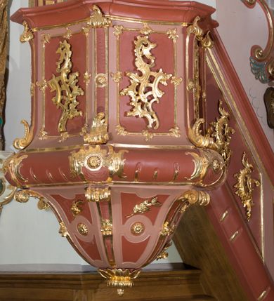 Zdjęcie nr 4: Ambona przyścienna, wisząca ustawiona przy północnej ścianie, dostępna jednobiegowymi schodami z balustradą od strony prezbiterium. Kosz na planie sześcioboku u dołu z ćwierćwałkiem, zdobionym na narożach dekoracją rokokową; u góry zamknięty profilowanym gzymsem. Ścianki artykułowane spływami wolutowymi o karbowanych trzonach. W polach prostokątne płyciny mieszczące płaskorzeźbione rocaille. Podwieszenie w formie stożka o wklęsło-wypukłych bokach, zakończonego płaskorzeźbioną rozetą. Ścianki flankowane w narożach analogicznie jak kosz, w polach trapezoidalne płyciny, przerwane pośrodku okręgiem z rozetką. Zaplecek prostokąty ujęty płaskim obramieniem o zgeometryzowanych wolutach w dolnych narożach oraz rocaille&#039;m w górnych. Pośrodku płaskorzeźbione przedstawienie św. Jana Chrzciciela na tle pejzażu. Baldachim powtarzający plan kosza, zwieńczony wazonem ustawionym na sześciu spiętych kabłąkach. W podniebiu gołębica Ducha Świętego na tle promienistej glorii, na brzegach zawieszony lambrekin. Balustrada schodów pełna, od dołu ujęta półwałkiem, zamknięta profilowanym gzymsem, w polu dwie  płyciny w kształcie rombów, w których umieszczone są rocaille. Balustrada zakończona filarkiem, na którym ustawiony jest wazonik. Całość struktury ambony polichromowana w odcieniach brązu, płaskorzeźba wielobarwna, profilowania i detal architektoniczny pozłacany, podniebienie baldachimu malowane na niebiesko, lambrekin posrebrzany.

