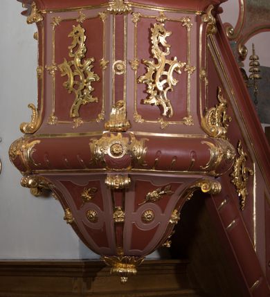 Zdjęcie nr 2: Ambona przyścienna, wisząca ustawiona przy północnej ścianie, dostępna jednobiegowymi schodami z balustradą od strony prezbiterium. Kosz na planie sześcioboku u dołu z ćwierćwałkiem, zdobionym na narożach dekoracją rokokową; u góry zamknięty profilowanym gzymsem. Ścianki artykułowane spływami wolutowymi o karbowanych trzonach. W polach prostokątne płyciny mieszczące płaskorzeźbione rocaille. Podwieszenie w formie stożka o wklęsło-wypukłych bokach, zakończonego płaskorzeźbioną rozetą. Ścianki flankowane w narożach analogicznie jak kosz, w polach trapezoidalne płyciny, przerwane pośrodku okręgiem z rozetką. Zaplecek prostokąty ujęty płaskim obramieniem o zgeometryzowanych wolutach w dolnych narożach oraz rocaille'm w górnych. Pośrodku płaskorzeźbione przedstawienie św. Jana Chrzciciela na tle pejzażu. Baldachim powtarzający plan kosza, zwieńczony wazonem ustawionym na sześciu spiętych kabłąkach. W podniebiu gołębica Ducha Świętego na tle promienistej glorii, na brzegach zawieszony lambrekin. Balustrada schodów pełna, od dołu ujęta półwałkiem, zamknięta profilowanym gzymsem, w polu dwie  płyciny w kształcie rombów, w których umieszczone są rocaille. Balustrada zakończona filarkiem, na którym ustawiony jest wazonik. Całość struktury ambony polichromowana w odcieniach brązu, płaskorzeźba wielobarwna, profilowania i detal architektoniczny pozłacany, podniebienie baldachimu malowane na niebiesko, lambrekin posrebrzany.

