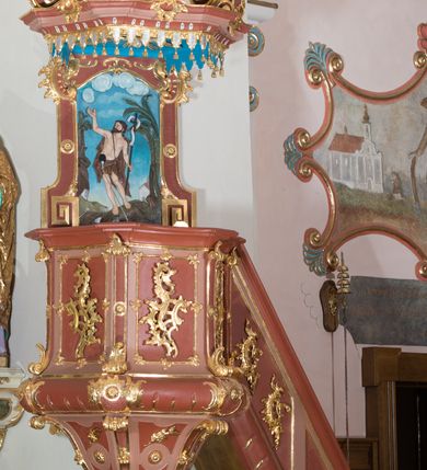Zdjęcie nr 5: Ambona przyścienna, wisząca ustawiona przy północnej ścianie, dostępna jednobiegowymi schodami z balustradą od strony prezbiterium. Kosz na planie sześcioboku u dołu z ćwierćwałkiem, zdobionym na narożach dekoracją rokokową; u góry zamknięty profilowanym gzymsem. Ścianki artykułowane spływami wolutowymi o karbowanych trzonach. W polach prostokątne płyciny mieszczące płaskorzeźbione rocaille. Podwieszenie w formie stożka o wklęsło-wypukłych bokach, zakończonego płaskorzeźbioną rozetą. Ścianki flankowane w narożach analogicznie jak kosz, w polach trapezoidalne płyciny, przerwane pośrodku okręgiem z rozetką. Zaplecek prostokąty ujęty płaskim obramieniem o zgeometryzowanych wolutach w dolnych narożach oraz rocaille'm w górnych. Pośrodku płaskorzeźbione przedstawienie św. Jana Chrzciciela na tle pejzażu. Baldachim powtarzający plan kosza, zwieńczony wazonem ustawionym na sześciu spiętych kabłąkach. W podniebiu gołębica Ducha Świętego na tle promienistej glorii, na brzegach zawieszony lambrekin. Balustrada schodów pełna, od dołu ujęta półwałkiem, zamknięta profilowanym gzymsem, w polu dwie  płyciny w kształcie rombów, w których umieszczone są rocaille. Balustrada zakończona filarkiem, na którym ustawiony jest wazonik. Całość struktury ambony polichromowana w odcieniach brązu, płaskorzeźba wielobarwna, profilowania i detal architektoniczny pozłacany, podniebienie baldachimu malowane na niebiesko, lambrekin posrebrzany.

