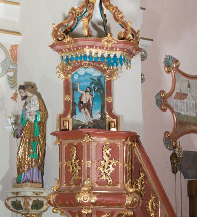 Zdjęcie nr 1: Ambona przyścienna, wisząca ustawiona przy północnej ścianie, dostępna jednobiegowymi schodami z balustradą od strony prezbiterium. Kosz na planie sześcioboku u dołu z ćwierćwałkiem, zdobionym na narożach dekoracją rokokową; u góry zamknięty profilowanym gzymsem. Ścianki artykułowane spływami wolutowymi o karbowanych trzonach. W polach prostokątne płyciny mieszczące płaskorzeźbione rocaille. Podwieszenie w formie stożka o wklęsło-wypukłych bokach, zakończonego płaskorzeźbioną rozetą. Ścianki flankowane w narożach analogicznie jak kosz, w polach trapezoidalne płyciny, przerwane pośrodku okręgiem z rozetką. Zaplecek prostokąty ujęty płaskim obramieniem o zgeometryzowanych wolutach w dolnych narożach oraz rocaille&#039;m w górnych. Pośrodku płaskorzeźbione przedstawienie św. Jana Chrzciciela na tle pejzażu. Baldachim powtarzający plan kosza, zwieńczony wazonem ustawionym na sześciu spiętych kabłąkach. W podniebiu gołębica Ducha Świętego na tle promienistej glorii, na brzegach zawieszony lambrekin. Balustrada schodów pełna, od dołu ujęta półwałkiem, zamknięta profilowanym gzymsem, w polu dwie  płyciny w kształcie rombów, w których umieszczone są rocaille. Balustrada zakończona filarkiem, na którym ustawiony jest wazonik. Całość struktury ambony polichromowana w odcieniach brązu, płaskorzeźba wielobarwna, profilowania i detal architektoniczny pozłacany, podniebienie baldachimu malowane na niebiesko, lambrekin posrebrzany.

