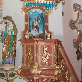 Zdjęcie nr 1: Ambona przyścienna, wisząca ustawiona przy północnej ścianie, dostępna jednobiegowymi schodami z balustradą od strony prezbiterium. Kosz na planie sześcioboku u dołu z ćwierćwałkiem, zdobionym na narożach dekoracją rokokową; u góry zamknięty profilowanym gzymsem. Ścianki artykułowane spływami wolutowymi o karbowanych trzonach. W polach prostokątne płyciny mieszczące płaskorzeźbione rocaille. Podwieszenie w formie stożka o wklęsło-wypukłych bokach, zakończonego płaskorzeźbioną rozetą. Ścianki flankowane w narożach analogicznie jak kosz, w polach trapezoidalne płyciny, przerwane pośrodku okręgiem z rozetką. Zaplecek prostokąty ujęty płaskim obramieniem o zgeometryzowanych wolutach w dolnych narożach oraz rocaille'm w górnych. Pośrodku płaskorzeźbione przedstawienie św. Jana Chrzciciela na tle pejzażu. Baldachim powtarzający plan kosza, zwieńczony wazonem ustawionym na sześciu spiętych kabłąkach. W podniebiu gołębica Ducha Świętego na tle promienistej glorii, na brzegach zawieszony lambrekin. Balustrada schodów pełna, od dołu ujęta półwałkiem, zamknięta profilowanym gzymsem, w polu dwie  płyciny w kształcie rombów, w których umieszczone są rocaille. Balustrada zakończona filarkiem, na którym ustawiony jest wazonik. Całość struktury ambony polichromowana w odcieniach brązu, płaskorzeźba wielobarwna, profilowania i detal architektoniczny pozłacany, podniebienie baldachimu malowane na niebiesko, lambrekin posrebrzany.

