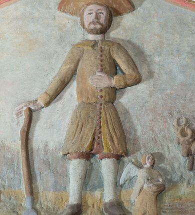 Zdjęcie nr 3: Płaskorzeźba ujęta w fantazyjną ramę utworzoną z zawijających się do środka wolut w kształcie litery „C” spiętych palmetami. Pośrodku ukazany św. Izydor Oracz stojący frontalnie w lekkim rozkroku z prawą ręką wspartą o laskę, lewą złożoną na piersi. Twarz szeroka, okolona krótką brodą. Święty ubrany jest w krótkie, czerwone spodnie, sukmanę sięgająca kolan oraz kapelusz, na stopach ma pełne, brązowe buty. Po prawej stronie kompozycji, u stóp  świętego  klęczący anioł. W tle pejzaż z bryłą kościoła w Podwilku i klęczącą przed nim postacią ukazaną po lewej stronie, natomiast po prawej pług zaprzężony do dwóch wołów, prowadzony przez anioła. Polichromia w odcieniach pastelowych, tunika i kapelusz świętego brązowy, kościół biały nakryty czerwonym dachem. Obramienie polichromii malowane na czerwono, pośrodku złocone, palmety malowane na niebiesko i złocone. Poniżej płaskorzeźby pole w kształcie leżącego prostokąta o wklęsłych narożach z napisem „Labore praeCIpVe pIIs eXpenIs sVbDlicae pLebIs hVIVs / paroChIae s VbViLkensIs haeC saCra aeDes feLICIter / sVrreXIt”.
