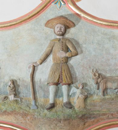 Zdjęcie nr 1: Płaskorzeźba ujęta w fantazyjną ramę utworzoną z zawijających się do środka wolut w kształcie litery „C” spiętych palmetami. Pośrodku ukazany św. Izydor Oracz stojący frontalnie w lekkim rozkroku z prawą ręką wspartą o laskę, lewą złożoną na piersi. Twarz szeroka, okolona krótką brodą. Święty ubrany jest w krótkie, czerwone spodnie, sukmanę sięgająca kolan oraz kapelusz, na stopach ma pełne, brązowe buty. Po prawej stronie kompozycji, u stóp  świętego  klęczący anioł. W tle pejzaż z bryłą kościoła w Podwilku i klęczącą przed nim postacią ukazaną po lewej stronie, natomiast po prawej pług zaprzężony do dwóch wołów, prowadzony przez anioła. Polichromia w odcieniach pastelowych, tunika i kapelusz świętego brązowy, kościół biały nakryty czerwonym dachem. Obramienie polichromii malowane na czerwono, pośrodku złocone, palmety malowane na niebiesko i złocone. Poniżej płaskorzeźby pole w kształcie leżącego prostokąta o wklęsłych narożach z napisem „Labore praeCIpVe pIIs eXpenIs sVbDlicae pLebIs hVIVs / paroChIae s VbViLkensIs haeC saCra aeDes feLICIter / sVrreXIt”.
