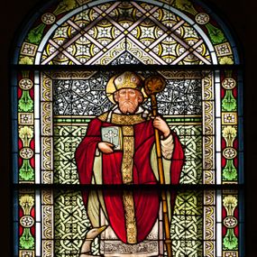 Zdjęcie nr 1: W centrum kompozycji całopostaciowe, frontalne przedstawienie stojącego św. Marcina w stroju biskupim: jasnoróżowej albie, białej komży, białym humerale oraz czerwonym ornacie z zieloną podszewką i złoconą pretekstą. Na głowie ma założoną infułę, a wokół niej złoty i kolisty nimb. W prawej dłoni trzyma księgę, przyciskając ją do piersi, a w lewej pastorał. Twarz szeroka z długim i wąskim nosem, małymi ustami, okolona średniej długości jasną brodą oraz krótkimi włosami. Przy prawej nodze świętego stoi biała gęś W tle ornamentalna tkanina z frędzlami u dołu. Całość ujęta półkolistą arkadą utworzoną z ułożonych naprzemiennie rozet oraz palmet. W dolnej części kompozycji prostokątne pole z inskrypcjami „Św(ięty) MARCIN / PALKA JOZSEF BUDAPESZT” oraz „RENOWACJA 1999 / PRACOWNIA WITRAŻU / GUSTAW KRAKÓW”.