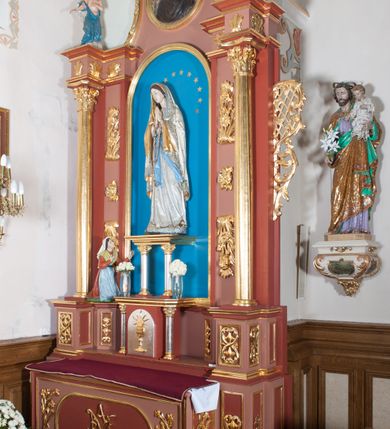 Zdjęcie nr 8: Nastawa ołtarzowa przyścienna, architektoniczna, jednoosiowa, jednokondygnacyjna ze zwieńczeniem. Ołtarz skrzyniowy, prostopadłościenny z kamiennym portatylem w drewnianej ramie. Antepedium drewniane, zdobione płyciną, w której umieszczony jest monogram Matki Boskiej. Po bokach płaskorzeźbione gałązki kwiatów. Tabernakulum w formie prostopadłościennej szafki flankowanej parą wolnostojących kolumienek o korynckich kapitelach, podtrzymujących profilowany gzyms płyty nakrywającej całość obiektu. Drzwiczki w formie zbliżonej do kwadratu, zamknięte półkoliście, zdobione płaskorzeźbioną dekoracją ukazującą kielich z hostią. 

Retabulum ustawione na wysokim, zdwojonym, uskokowym cokole. Nastawa ujęta parą kolumn korynckich. Kolumny wspierają przerwane, wyłamane ponad nimi belkowanie z wysuniętym gzymsem. Pola środkowe w formie prostokątnej wnęki zamkniętej półkoliście, ujętej profilowaną ramą. Wewnątrz pola rzeźba Matki Boskiej ustawiona na wsporniku, wspartym na dwóch kolumienkach. Po lewej stronie rzeźba św. Bernadetty z Soubirous. Tło malowane na kolor błękitny. W partii fryzu motyw stylizowanej palmety. Po bokach nastawy uszy z liści akantu oplatające kratkę regencyjną. W pionowych polach flankujących główną scenę ołtarza ornamentyka roślinna liści akantu z rozetami, pomiędzy nimi motyw kwiatu róży. 

Zwieńczenie o formie wklęsło-wypukłej, ujęte dekoracyjnymi spływami o formach roślinnych. W polu płaskorzeźba przedstawiająca popiersie św. Maksymiliana Marii Kolbego. Po bokach zwieńczenia dwie rzeźby aniołów. Polichromia całości struktury ołtarza w odcieniach brązu, profilowania, detal architektoniczny i ornamentyka złocone i srebrzone.  





