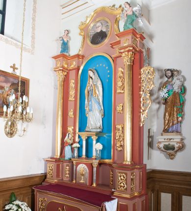 Zdjęcie nr 1: Nastawa ołtarzowa przyścienna, architektoniczna, jednoosiowa, jednokondygnacyjna ze zwieńczeniem. Ołtarz skrzyniowy, prostopadłościenny z kamiennym portatylem w drewnianej ramie. Antepedium drewniane, zdobione płyciną, w której umieszczony jest monogram Matki Boskiej. Po bokach płaskorzeźbione gałązki kwiatów. Tabernakulum w formie prostopadłościennej szafki flankowanej parą wolnostojących kolumienek o korynckich kapitelach, podtrzymujących profilowany gzyms płyty nakrywającej całość obiektu. Drzwiczki w formie zbliżonej do kwadratu, zamknięte półkoliście, zdobione płaskorzeźbioną dekoracją ukazującą kielich z hostią. 

Retabulum ustawione na wysokim, zdwojonym, uskokowym cokole. Nastawa ujęta parą kolumn korynckich. Kolumny wspierają przerwane, wyłamane ponad nimi belkowanie z wysuniętym gzymsem. Pola środkowe w formie prostokątnej wnęki zamkniętej półkoliście, ujętej profilowaną ramą. Wewnątrz pola rzeźba Matki Boskiej ustawiona na wsporniku, wspartym na dwóch kolumienkach. Po lewej stronie rzeźba św. Bernadetty z Soubirous. Tło malowane na kolor błękitny. W partii fryzu motyw stylizowanej palmety. Po bokach nastawy uszy z liści akantu oplatające kratkę regencyjną. W pionowych polach flankujących główną scenę ołtarza ornamentyka roślinna liści akantu z rozetami, pomiędzy nimi motyw kwiatu róży. 

Zwieńczenie o formie wklęsło-wypukłej, ujęte dekoracyjnymi spływami o formach roślinnych. W polu płaskorzeźba przedstawiająca popiersie św. Maksymiliana Marii Kolbego. Po bokach zwieńczenia dwie rzeźby aniołów. Polichromia całości struktury ołtarza w odcieniach brązu, profilowania, detal architektoniczny i ornamentyka złocone i srebrzone.  





