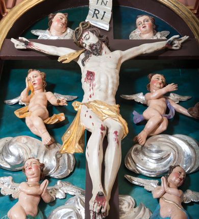 Zdjęcie nr 9: Pełnoplastyczna, polichromowana figura Chrystusa w typie Cristo morte przybitego trzema gwoździami do  dużego, brązowego krzyża. Chrystus ukazany w statycznej pozie, w mocnym zwisie z ugiętymi w kolanach nogami. Twarz podłużna z długim nosem, okolona krótką brodą, która na końcu zawija się w dwa pukle. Włosy długie, ciemnobrązowe opadające na prawe ramię i plecy. Sylwetka szczupła o wyraźnie podkreślonej linii żeber z guzowatą klatką piersiową; na głowie, kolanach, dłoniach, stopach i boku ślady krwi.  Perizonium przewiązane na prawym boku ze zwisem wzdłuż prawego uda, złocone. Na głowie szeroka korona cierniowa, wokół niej niekompletny nimb krzyżowy z jedną wiązką promieni. Kolorystyka ciała utrzymana w odcieniach bladego  beżu. U góry pionowej belki posrebrzany titulus w formie pionowej pogiętej banderoli z napisem: „IN / RI”. Krucyfiks otaczają cztery aniołki na obłokach o teatralnych gestach, nad krzyżem dwie uskrzydlone główki anielskie. Tło malowane na kolor ciemnoniebieski. Polichromia w odsłoniętych partiach ciała naturalistyczna, detale złocone i srebrzone.