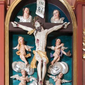 Zdjęcie nr 1: Pełnoplastyczna, polichromowana figura Chrystusa w typie Cristo morte przybitego trzema gwoździami do  dużego, brązowego krzyża. Chrystus ukazany w statycznej pozie, w mocnym zwisie z ugiętymi w kolanach nogami. Twarz podłużna z długim nosem, okolona krótką brodą, która na końcu zawija się w dwa pukle. Włosy długie, ciemnobrązowe opadające na prawe ramię i plecy. Sylwetka szczupła o wyraźnie podkreślonej linii żeber z guzowatą klatką piersiową; na głowie, kolanach, dłoniach, stopach i boku ślady krwi.  Perizonium przewiązane na prawym boku ze zwisem wzdłuż prawego uda, złocone. Na głowie szeroka korona cierniowa, wokół niej niekompletny nimb krzyżowy z jedną wiązką promieni. Kolorystyka ciała utrzymana w odcieniach bladego  beżu. U góry pionowej belki posrebrzany titulus w formie pionowej pogiętej banderoli z napisem: „IN / RI”. Krucyfiks otaczają cztery aniołki na obłokach o teatralnych gestach, nad krzyżem dwie uskrzydlone główki anielskie. Tło malowane na kolor ciemnoniebieski. Polichromia w odsłoniętych partiach ciała naturalistyczna, detale złocone i srebrzone.