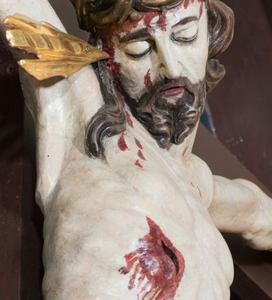 Zdjęcie nr 10: Pełnoplastyczna, polichromowana figura Chrystusa w typie Cristo morte przybitego trzema gwoździami do  dużego, brązowego krzyża. Chrystus ukazany w statycznej pozie, w mocnym zwisie z ugiętymi w kolanach nogami. Twarz podłużna z długim nosem, okolona krótką brodą, która na końcu zawija się w dwa pukle. Włosy długie, ciemnobrązowe opadające na prawe ramię i plecy. Sylwetka szczupła o wyraźnie podkreślonej linii żeber z guzowatą klatką piersiową; na głowie, kolanach, dłoniach, stopach i boku ślady krwi.  Perizonium przewiązane na prawym boku ze zwisem wzdłuż prawego uda, złocone. Na głowie szeroka korona cierniowa, wokół niej niekompletny nimb krzyżowy z jedną wiązką promieni. Kolorystyka ciała utrzymana w odcieniach bladego  beżu. U góry pionowej belki posrebrzany titulus w formie pionowej pogiętej banderoli z napisem: „IN / RI”. Krucyfiks otaczają cztery aniołki na obłokach o teatralnych gestach, nad krzyżem dwie uskrzydlone główki anielskie. Tło malowane na kolor ciemnoniebieski. Polichromia w odsłoniętych partiach ciała naturalistyczna, detale złocone i srebrzone.