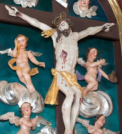 Zdjęcie nr 2: Pełnoplastyczna, polichromowana figura Chrystusa w typie Cristo morte przybitego trzema gwoździami do  dużego, brązowego krzyża. Chrystus ukazany w statycznej pozie, w mocnym zwisie z ugiętymi w kolanach nogami. Twarz podłużna z długim nosem, okolona krótką brodą, która na końcu zawija się w dwa pukle. Włosy długie, ciemnobrązowe opadające na prawe ramię i plecy. Sylwetka szczupła o wyraźnie podkreślonej linii żeber z guzowatą klatką piersiową; na głowie, kolanach, dłoniach, stopach i boku ślady krwi.  Perizonium przewiązane na prawym boku ze zwisem wzdłuż prawego uda, złocone. Na głowie szeroka korona cierniowa, wokół niej niekompletny nimb krzyżowy z jedną wiązką promieni. Kolorystyka ciała utrzymana w odcieniach bladego  beżu. U góry pionowej belki posrebrzany titulus w formie pionowej pogiętej banderoli z napisem: „IN / RI”. Krucyfiks otaczają cztery aniołki na obłokach o teatralnych gestach, nad krzyżem dwie uskrzydlone główki anielskie. Tło malowane na kolor ciemnoniebieski. Polichromia w odsłoniętych partiach ciała naturalistyczna, detale złocone i srebrzone.