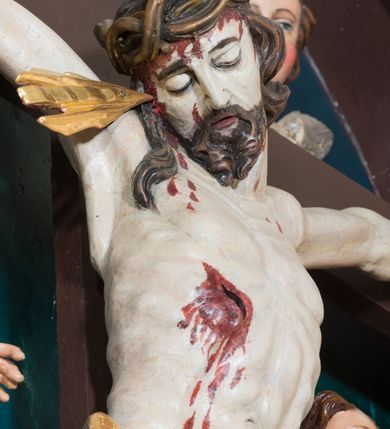 Zdjęcie nr 11: Pełnoplastyczna, polichromowana figura Chrystusa w typie Cristo morte przybitego trzema gwoździami do  dużego, brązowego krzyża. Chrystus ukazany w statycznej pozie, w mocnym zwisie z ugiętymi w kolanach nogami. Twarz podłużna z długim nosem, okolona krótką brodą, która na końcu zawija się w dwa pukle. Włosy długie, ciemnobrązowe opadające na prawe ramię i plecy. Sylwetka szczupła o wyraźnie podkreślonej linii żeber z guzowatą klatką piersiową; na głowie, kolanach, dłoniach, stopach i boku ślady krwi.  Perizonium przewiązane na prawym boku ze zwisem wzdłuż prawego uda, złocone. Na głowie szeroka korona cierniowa, wokół niej niekompletny nimb krzyżowy z jedną wiązką promieni. Kolorystyka ciała utrzymana w odcieniach bladego  beżu. U góry pionowej belki posrebrzany titulus w formie pionowej pogiętej banderoli z napisem: „IN / RI”. Krucyfiks otaczają cztery aniołki na obłokach o teatralnych gestach, nad krzyżem dwie uskrzydlone główki anielskie. Tło malowane na kolor ciemnoniebieski. Polichromia w odsłoniętych partiach ciała naturalistyczna, detale złocone i srebrzone.