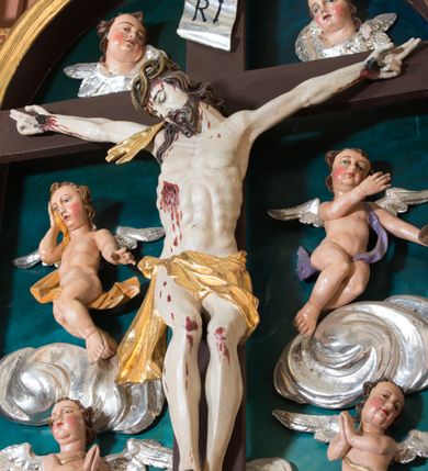 Zdjęcie nr 12: Pełnoplastyczna, polichromowana figura Chrystusa w typie Cristo morte przybitego trzema gwoździami do  dużego, brązowego krzyża. Chrystus ukazany w statycznej pozie, w mocnym zwisie z ugiętymi w kolanach nogami. Twarz podłużna z długim nosem, okolona krótką brodą, która na końcu zawija się w dwa pukle. Włosy długie, ciemnobrązowe opadające na prawe ramię i plecy. Sylwetka szczupła o wyraźnie podkreślonej linii żeber z guzowatą klatką piersiową; na głowie, kolanach, dłoniach, stopach i boku ślady krwi.  Perizonium przewiązane na prawym boku ze zwisem wzdłuż prawego uda, złocone. Na głowie szeroka korona cierniowa, wokół niej niekompletny nimb krzyżowy z jedną wiązką promieni. Kolorystyka ciała utrzymana w odcieniach bladego  beżu. U góry pionowej belki posrebrzany titulus w formie pionowej pogiętej banderoli z napisem: „IN / RI”. Krucyfiks otaczają cztery aniołki na obłokach o teatralnych gestach, nad krzyżem dwie uskrzydlone główki anielskie. Tło malowane na kolor ciemnoniebieski. Polichromia w odsłoniętych partiach ciała naturalistyczna, detale złocone i srebrzone.
