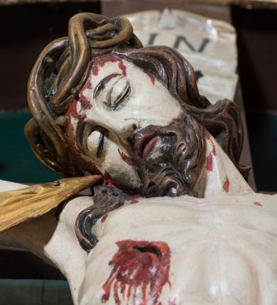 Zdjęcie nr 5: Pełnoplastyczna, polichromowana figura Chrystusa w typie Cristo morte przybitego trzema gwoździami do  dużego, brązowego krzyża. Chrystus ukazany w statycznej pozie, w mocnym zwisie z ugiętymi w kolanach nogami. Twarz podłużna z długim nosem, okolona krótką brodą, która na końcu zawija się w dwa pukle. Włosy długie, ciemnobrązowe opadające na prawe ramię i plecy. Sylwetka szczupła o wyraźnie podkreślonej linii żeber z guzowatą klatką piersiową; na głowie, kolanach, dłoniach, stopach i boku ślady krwi.  Perizonium przewiązane na prawym boku ze zwisem wzdłuż prawego uda, złocone. Na głowie szeroka korona cierniowa, wokół niej niekompletny nimb krzyżowy z jedną wiązką promieni. Kolorystyka ciała utrzymana w odcieniach bladego  beżu. U góry pionowej belki posrebrzany titulus w formie pionowej pogiętej banderoli z napisem: „IN / RI”. Krucyfiks otaczają cztery aniołki na obłokach o teatralnych gestach, nad krzyżem dwie uskrzydlone główki anielskie. Tło malowane na kolor ciemnoniebieski. Polichromia w odsłoniętych partiach ciała naturalistyczna, detale złocone i srebrzone.