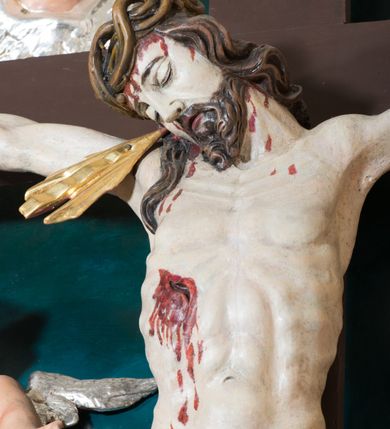 Zdjęcie nr 7: Pełnoplastyczna, polichromowana figura Chrystusa w typie Cristo morte przybitego trzema gwoździami do  dużego, brązowego krzyża. Chrystus ukazany w statycznej pozie, w mocnym zwisie z ugiętymi w kolanach nogami. Twarz podłużna z długim nosem, okolona krótką brodą, która na końcu zawija się w dwa pukle. Włosy długie, ciemnobrązowe opadające na prawe ramię i plecy. Sylwetka szczupła o wyraźnie podkreślonej linii żeber z guzowatą klatką piersiową; na głowie, kolanach, dłoniach, stopach i boku ślady krwi.  Perizonium przewiązane na prawym boku ze zwisem wzdłuż prawego uda, złocone. Na głowie szeroka korona cierniowa, wokół niej niekompletny nimb krzyżowy z jedną wiązką promieni. Kolorystyka ciała utrzymana w odcieniach bladego  beżu. U góry pionowej belki posrebrzany titulus w formie pionowej pogiętej banderoli z napisem: „IN / RI”. Krucyfiks otaczają cztery aniołki na obłokach o teatralnych gestach, nad krzyżem dwie uskrzydlone główki anielskie. Tło malowane na kolor ciemnoniebieski. Polichromia w odsłoniętych partiach ciała naturalistyczna, detale złocone i srebrzone.