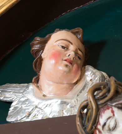 Zdjęcie nr 4: Pełnoplastyczna, polichromowana figura Chrystusa w typie Cristo morte przybitego trzema gwoździami do  dużego, brązowego krzyża. Chrystus ukazany w statycznej pozie, w mocnym zwisie z ugiętymi w kolanach nogami. Twarz podłużna z długim nosem, okolona krótką brodą, która na końcu zawija się w dwa pukle. Włosy długie, ciemnobrązowe opadające na prawe ramię i plecy. Sylwetka szczupła o wyraźnie podkreślonej linii żeber z guzowatą klatką piersiową; na głowie, kolanach, dłoniach, stopach i boku ślady krwi.  Perizonium przewiązane na prawym boku ze zwisem wzdłuż prawego uda, złocone. Na głowie szeroka korona cierniowa, wokół niej niekompletny nimb krzyżowy z jedną wiązką promieni. Kolorystyka ciała utrzymana w odcieniach bladego  beżu. U góry pionowej belki posrebrzany titulus w formie pionowej pogiętej banderoli z napisem: „IN / RI”. Krucyfiks otaczają cztery aniołki na obłokach o teatralnych gestach, nad krzyżem dwie uskrzydlone główki anielskie. Tło malowane na kolor ciemnoniebieski. Polichromia w odsłoniętych partiach ciała naturalistyczna, detale złocone i srebrzone.