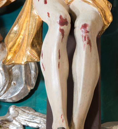 Zdjęcie nr 6: Pełnoplastyczna, polichromowana figura Chrystusa w typie Cristo morte przybitego trzema gwoździami do  dużego, brązowego krzyża. Chrystus ukazany w statycznej pozie, w mocnym zwisie z ugiętymi w kolanach nogami. Twarz podłużna z długim nosem, okolona krótką brodą, która na końcu zawija się w dwa pukle. Włosy długie, ciemnobrązowe opadające na prawe ramię i plecy. Sylwetka szczupła o wyraźnie podkreślonej linii żeber z guzowatą klatką piersiową; na głowie, kolanach, dłoniach, stopach i boku ślady krwi.  Perizonium przewiązane na prawym boku ze zwisem wzdłuż prawego uda, złocone. Na głowie szeroka korona cierniowa, wokół niej niekompletny nimb krzyżowy z jedną wiązką promieni. Kolorystyka ciała utrzymana w odcieniach bladego  beżu. U góry pionowej belki posrebrzany titulus w formie pionowej pogiętej banderoli z napisem: „IN / RI”. Krucyfiks otaczają cztery aniołki na obłokach o teatralnych gestach, nad krzyżem dwie uskrzydlone główki anielskie. Tło malowane na kolor ciemnoniebieski. Polichromia w odsłoniętych partiach ciała naturalistyczna, detale złocone i srebrzone.