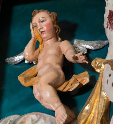 Zdjęcie nr 8: Pełnoplastyczna, polichromowana figura Chrystusa w typie Cristo morte przybitego trzema gwoździami do  dużego, brązowego krzyża. Chrystus ukazany w statycznej pozie, w mocnym zwisie z ugiętymi w kolanach nogami. Twarz podłużna z długim nosem, okolona krótką brodą, która na końcu zawija się w dwa pukle. Włosy długie, ciemnobrązowe opadające na prawe ramię i plecy. Sylwetka szczupła o wyraźnie podkreślonej linii żeber z guzowatą klatką piersiową; na głowie, kolanach, dłoniach, stopach i boku ślady krwi.  Perizonium przewiązane na prawym boku ze zwisem wzdłuż prawego uda, złocone. Na głowie szeroka korona cierniowa, wokół niej niekompletny nimb krzyżowy z jedną wiązką promieni. Kolorystyka ciała utrzymana w odcieniach bladego  beżu. U góry pionowej belki posrebrzany titulus w formie pionowej pogiętej banderoli z napisem: „IN / RI”. Krucyfiks otaczają cztery aniołki na obłokach o teatralnych gestach, nad krzyżem dwie uskrzydlone główki anielskie. Tło malowane na kolor ciemnoniebieski. Polichromia w odsłoniętych partiach ciała naturalistyczna, detale złocone i srebrzone.