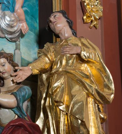 Zdjęcie nr 2: Rzeźba wolnostojąca, wydrążona, umieszczona  na srebrzonej podstawie imitującej kamień. Święta Maria Magdalena przedstawiona w ujęciu całopostaciowym, w pozycji klęczącej, zwrócona w trzech czwartych w prawo, oburącz obejmująca krzyż. Twarz pełna, oczy opuszczone w dół, przymknięte, nos i usta niewielkie. Karnacja jasna o zaróżowionych policzkach. Włosy długie, falowane, brązowe, opadające na plecy i prawe ramię. Ubrana jest w długą biało-błękitną suknię z szerokimi, podwiniętymi rękawami oraz czerwony płaszcz okrywający część pleców i lewą nogę. Polichromia w odsłoniętych partiach ciała naturalistyczna.