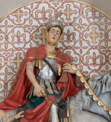 Zdjęcie nr 1: Rzeźba św. Marcina, drążona przedstawiająca świętego, który siedzi na koniu o szarej maści i ucina połę płaszcza biedakowi stojącemu po lewej stronie kompozycji. Święty zwrócony w lewo, ukazany w pozycji siedzącej, lewą dłonią trzyma uzdę konia, w prawej ma miecz. Twarz podłużna z długim nosem i przymkniętymi oczami, policzki silnie zarumienione. Ubrany jest w strój legionisty rzymskiego: pełną zbroję, hełm, napierśnik, ostrogi, zieloną tunikę oraz czerwony płaszcz spięty pod szyją złotą klamrę, ozdobiony na brzegu złoconą dekoracją roślinną. Z lewej, u dołu przedstawiony jest starszy mężczyzna, który siedzi na skale i dłońmi chwyta płaszcz św. Marcina. Ubrany jest w zieloną szatę ze srebrzoną lamówką, która osłania jego biodra. Twarz o rysach starszego mężczyzny, wychudzona, okolona długą i siwą brodą oraz rzadkimi włosami. Wnęka zdobiona krzyżami greckimi wpisanymi w czwórliście. Polichromia w odsłoniętych partiach ciała naturalistyczna, elementy uzbrojenia złocone i srebrzone.
