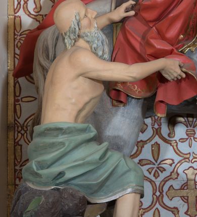 Zdjęcie nr 3: Rzeźba św. Marcina, drążona przedstawiająca świętego, który siedzi na koniu o szarej maści i ucina połę płaszcza biedakowi stojącemu po lewej stronie kompozycji. Święty zwrócony w lewo, ukazany w pozycji siedzącej, lewą dłonią trzyma uzdę konia, w prawej ma miecz. Twarz podłużna z długim nosem i przymkniętymi oczami, policzki silnie zarumienione. Ubrany jest w strój legionisty rzymskiego: pełną zbroję, hełm, napierśnik, ostrogi, zieloną tunikę oraz czerwony płaszcz spięty pod szyją złotą klamrę, ozdobiony na brzegu złoconą dekoracją roślinną. Z lewej, u dołu przedstawiony jest starszy mężczyzna, który siedzi na skale i dłońmi chwyta płaszcz św. Marcina. Ubrany jest w zieloną szatę ze srebrzoną lamówką, która osłania jego biodra. Twarz o rysach starszego mężczyzny, wychudzona, okolona długą i siwą brodą oraz rzadkimi włosami. Wnęka zdobiona krzyżami greckimi wpisanymi w czwórliście. Polichromia w odsłoniętych partiach ciała naturalistyczna, elementy uzbrojenia złocone i srebrzone.