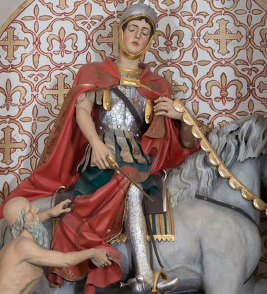 Zdjęcie nr 4: Rzeźba św. Marcina, drążona przedstawiająca świętego, który siedzi na koniu o szarej maści i ucina połę płaszcza biedakowi stojącemu po lewej stronie kompozycji. Święty zwrócony w lewo, ukazany w pozycji siedzącej, lewą dłonią trzyma uzdę konia, w prawej ma miecz. Twarz podłużna z długim nosem i przymkniętymi oczami, policzki silnie zarumienione. Ubrany jest w strój legionisty rzymskiego: pełną zbroję, hełm, napierśnik, ostrogi, zieloną tunikę oraz czerwony płaszcz spięty pod szyją złotą klamrę, ozdobiony na brzegu złoconą dekoracją roślinną. Z lewej, u dołu przedstawiony jest starszy mężczyzna, który siedzi na skale i dłońmi chwyta płaszcz św. Marcina. Ubrany jest w zieloną szatę ze srebrzoną lamówką, która osłania jego biodra. Twarz o rysach starszego mężczyzny, wychudzona, okolona długą i siwą brodą oraz rzadkimi włosami. Wnęka zdobiona krzyżami greckimi wpisanymi w czwórliście. Polichromia w odsłoniętych partiach ciała naturalistyczna, elementy uzbrojenia złocone i srebrzone.