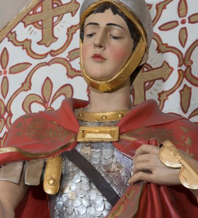 Zdjęcie nr 8: Rzeźba św. Marcina, drążona przedstawiająca świętego, który siedzi na koniu o szarej maści i ucina połę płaszcza biedakowi stojącemu po lewej stronie kompozycji. Święty zwrócony w lewo, ukazany w pozycji siedzącej, lewą dłonią trzyma uzdę konia, w prawej ma miecz. Twarz podłużna z długim nosem i przymkniętymi oczami, policzki silnie zarumienione. Ubrany jest w strój legionisty rzymskiego: pełną zbroję, hełm, napierśnik, ostrogi, zieloną tunikę oraz czerwony płaszcz spięty pod szyją złotą klamrę, ozdobiony na brzegu złoconą dekoracją roślinną. Z lewej, u dołu przedstawiony jest starszy mężczyzna, który siedzi na skale i dłońmi chwyta płaszcz św. Marcina. Ubrany jest w zieloną szatę ze srebrzoną lamówką, która osłania jego biodra. Twarz o rysach starszego mężczyzny, wychudzona, okolona długą i siwą brodą oraz rzadkimi włosami. Wnęka zdobiona krzyżami greckimi wpisanymi w czwórliście. Polichromia w odsłoniętych partiach ciała naturalistyczna, elementy uzbrojenia złocone i srebrzone.