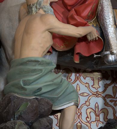 Zdjęcie nr 9: Rzeźba św. Marcina, drążona przedstawiająca świętego, który siedzi na koniu o szarej maści i ucina połę płaszcza biedakowi stojącemu po lewej stronie kompozycji. Święty zwrócony w lewo, ukazany w pozycji siedzącej, lewą dłonią trzyma uzdę konia, w prawej ma miecz. Twarz podłużna z długim nosem i przymkniętymi oczami, policzki silnie zarumienione. Ubrany jest w strój legionisty rzymskiego: pełną zbroję, hełm, napierśnik, ostrogi, zieloną tunikę oraz czerwony płaszcz spięty pod szyją złotą klamrę, ozdobiony na brzegu złoconą dekoracją roślinną. Z lewej, u dołu przedstawiony jest starszy mężczyzna, który siedzi na skale i dłońmi chwyta płaszcz św. Marcina. Ubrany jest w zieloną szatę ze srebrzoną lamówką, która osłania jego biodra. Twarz o rysach starszego mężczyzny, wychudzona, okolona długą i siwą brodą oraz rzadkimi włosami. Wnęka zdobiona krzyżami greckimi wpisanymi w czwórliście. Polichromia w odsłoniętych partiach ciała naturalistyczna, elementy uzbrojenia złocone i srebrzone.