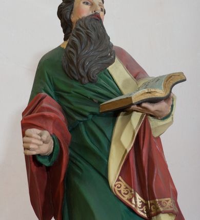 Zdjęcie nr 4: Rzeźba drążona, przedstawiająca św. Pawła. Święty ustawiony na niskim, brązowym cokole, frontalnie, w kontrapoście, o esowato wygiętej sylwetce, z prawą nogą ugiętą w kolanie, głową zwróconą w lewo. W lewej dłoni trzyma otwartą księgę, natomiast prawą ma wysuniętą do przodu i ułożoną do niezachowanego atrybutu (miecza). Twarz owalna z zarumienionymi policzkami z dużym nosem, okolona ciemnobrązową, gęstą i długą brodą. Włosy średniej długości, ciemnobrązowe, zasłaniają uszy. Ubrany jest w długą w zieloną suknię oraz czerwony płaszcz z beżową podszewką, założony na lewe ramię i przerzucony jedną połą przez prawe przedramię. Płaszcz na brzegach zdobiony jest złoconą dekoracją ornamentalną, a suknia u dołu dekorowana ornamentem złożonym ze złączonych trójliści. Polichromia w odsłoniętych partiach ciała naturalistyczna, detale złocone. 