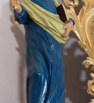 Zdjęcie nr 3: Rzeźba drążona, przedstawiająca św. Piotra. Święty ustawiony na niskim cokole, w delikatnym kontrapoście, z prawą nogą ugiętą w kolanie, z rozłożonymi na boki stopami. W prawej dłoni trzyma dwa skrzyżowane klucze, a w lewej księgę w brązowej oprawie. Twarz podłużna, okolona siwą brodą z długim nosem i małymi ustami. Włosy siwe, zasłaniające uszy, głowa ujęta do połowy łysiną, nad czołem kosmyk siwych włosów. Święty jest ubrany w długą, niebieską suknię z kołnierzykiem, u dołu zdobioną złoconym pasem ornamentu składającego się ze złączonych trójliści. Na suknię ma założony zwinięty jasnozielony płaszcz. Polichromia w odsłoniętych partiach ciała naturalistyczna, detale i klucze złocone.
